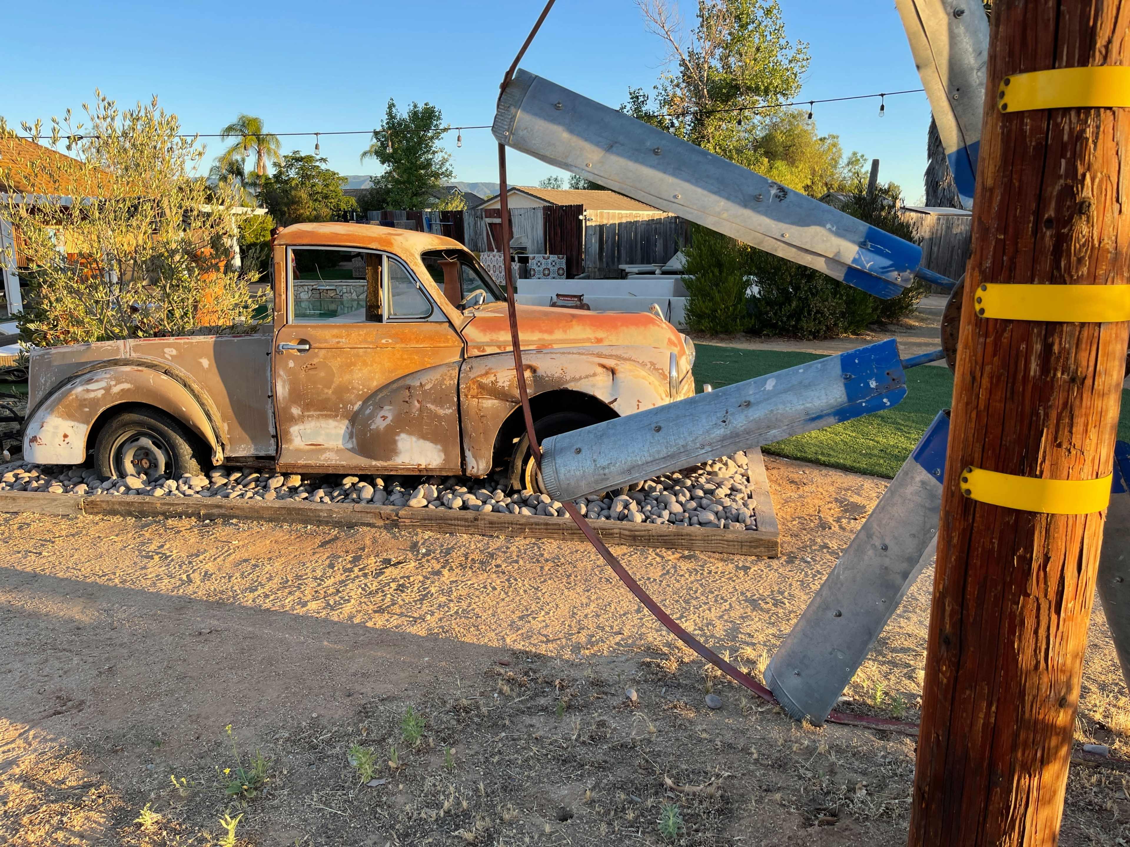 An old, rusty pickup truck is parked beside a wooden pole with metal supports, situated in a gravel area of a yard.