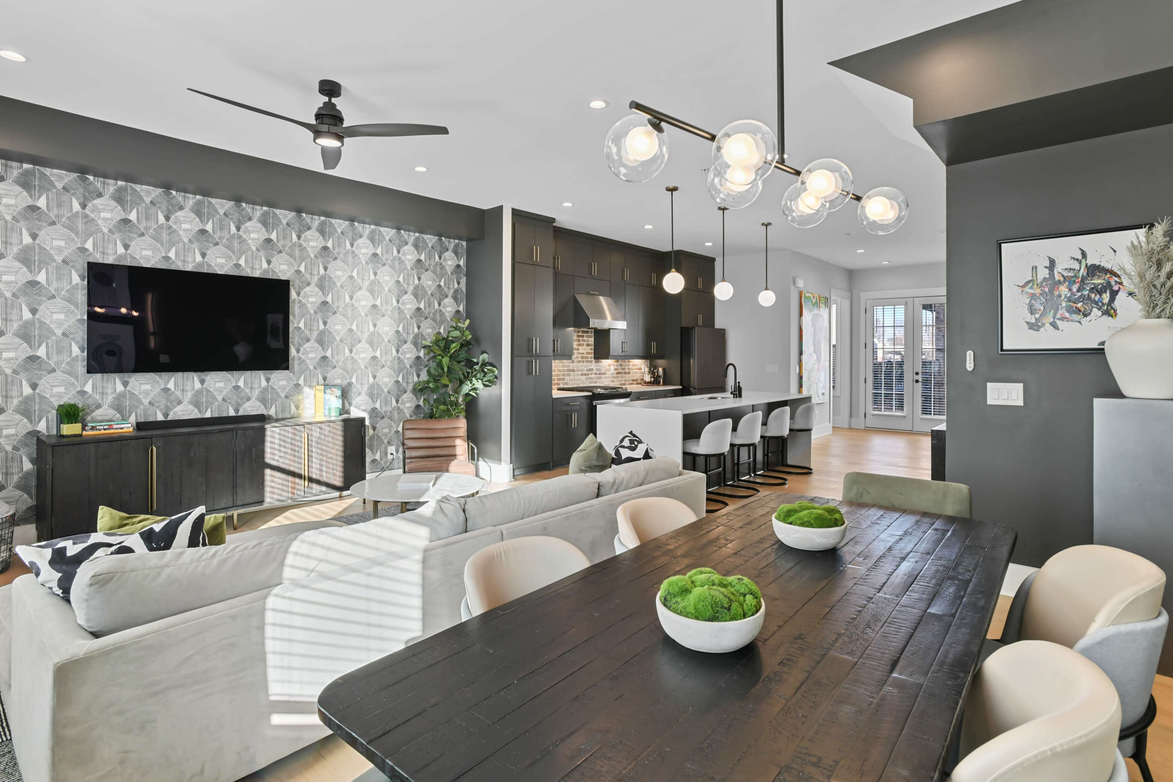 A modern open-concept living space features a dining table with bowls of greenery, a sectional sofa facing a wall-mounted TV, and a kitchen area with bar seating.