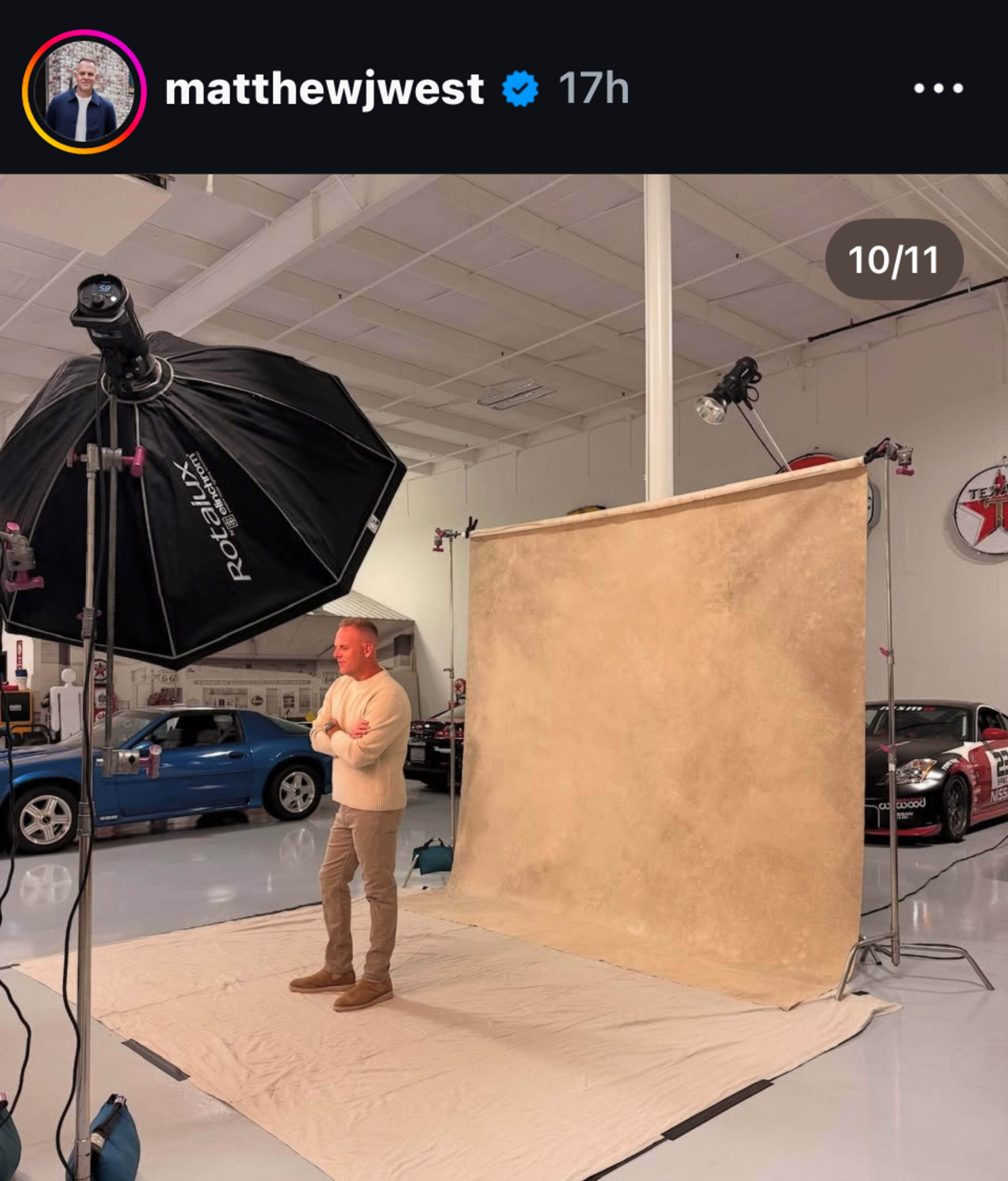 A man stands on a light-colored backdrop in a photography studio with a large softbox light overhead and sports cars visible in the background.