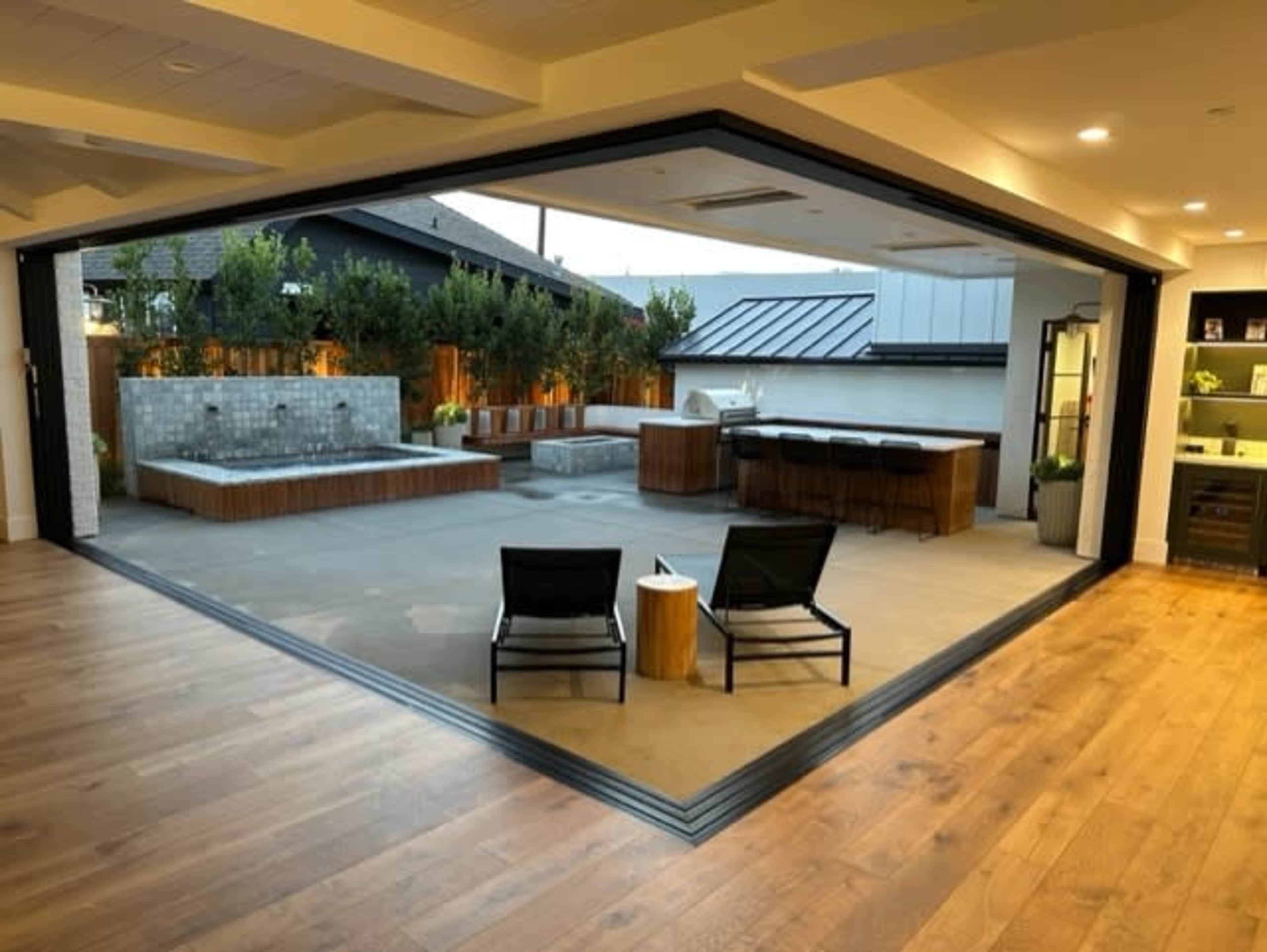 The image shows an interior space with large sliding glass doors that open to an outdoor area featuring a hot tub, seating, and a barbecue station.