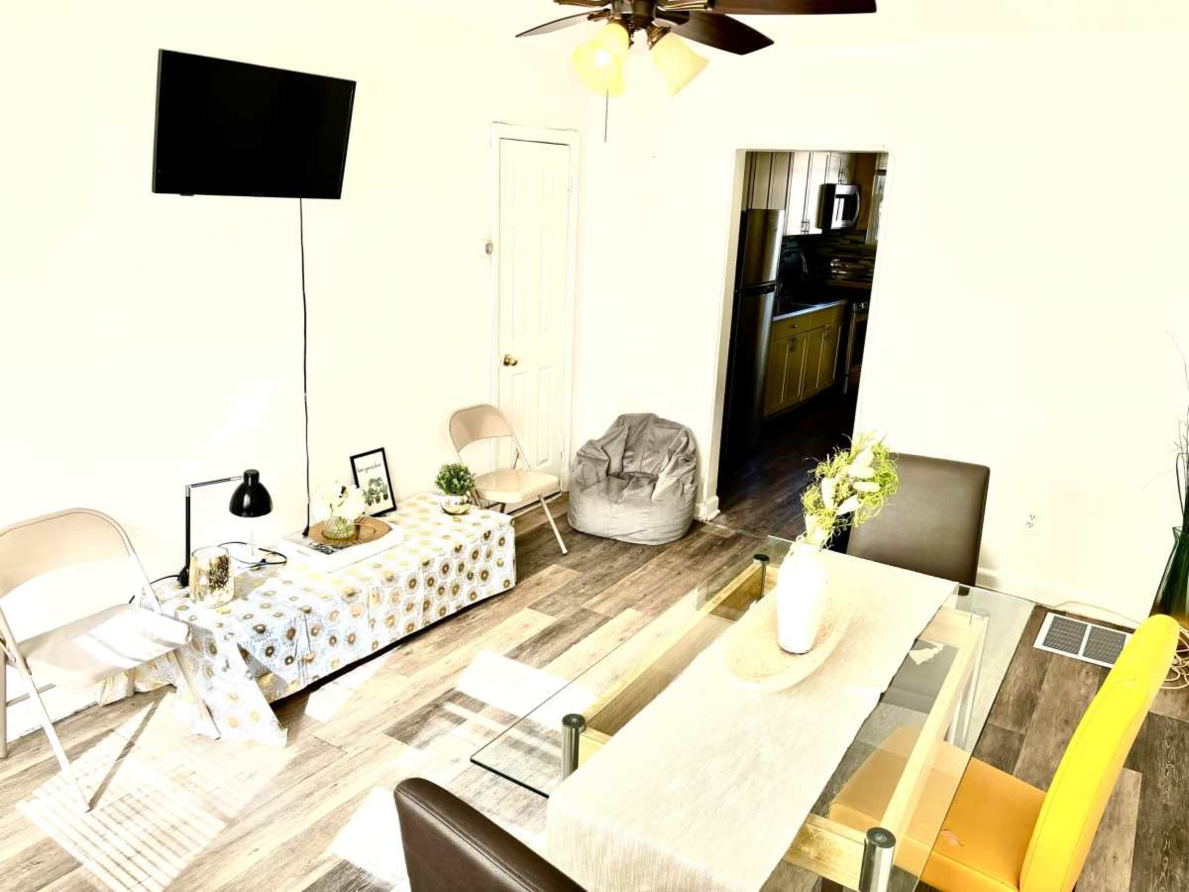 The image shows a living area with a glass dining table, two chairs, a beanbag, and a television mounted on the wall.