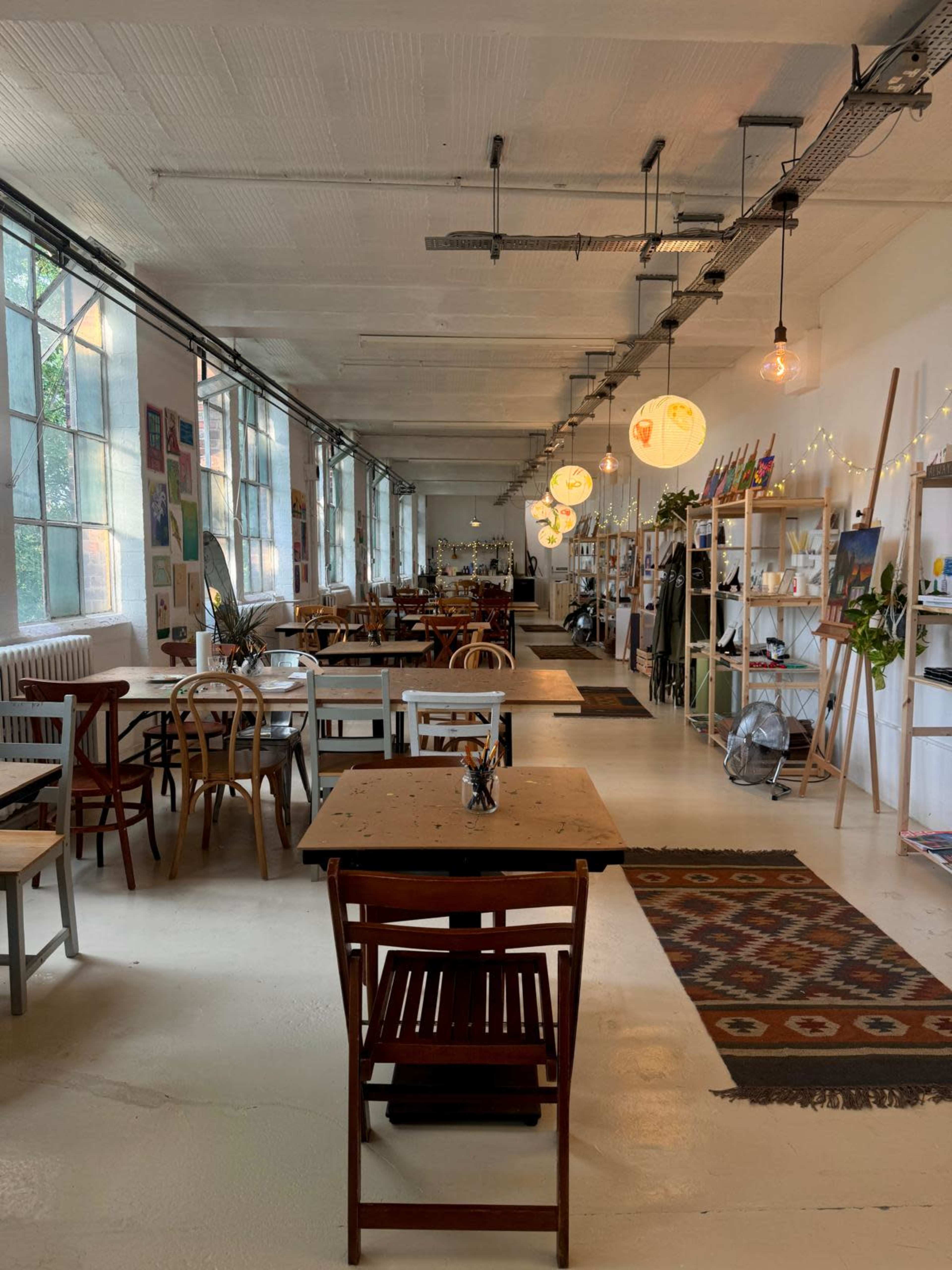 The image shows a spacious café with wooden tables and chairs, large windows, and decorative lighting fixtures hanging from the ceiling.