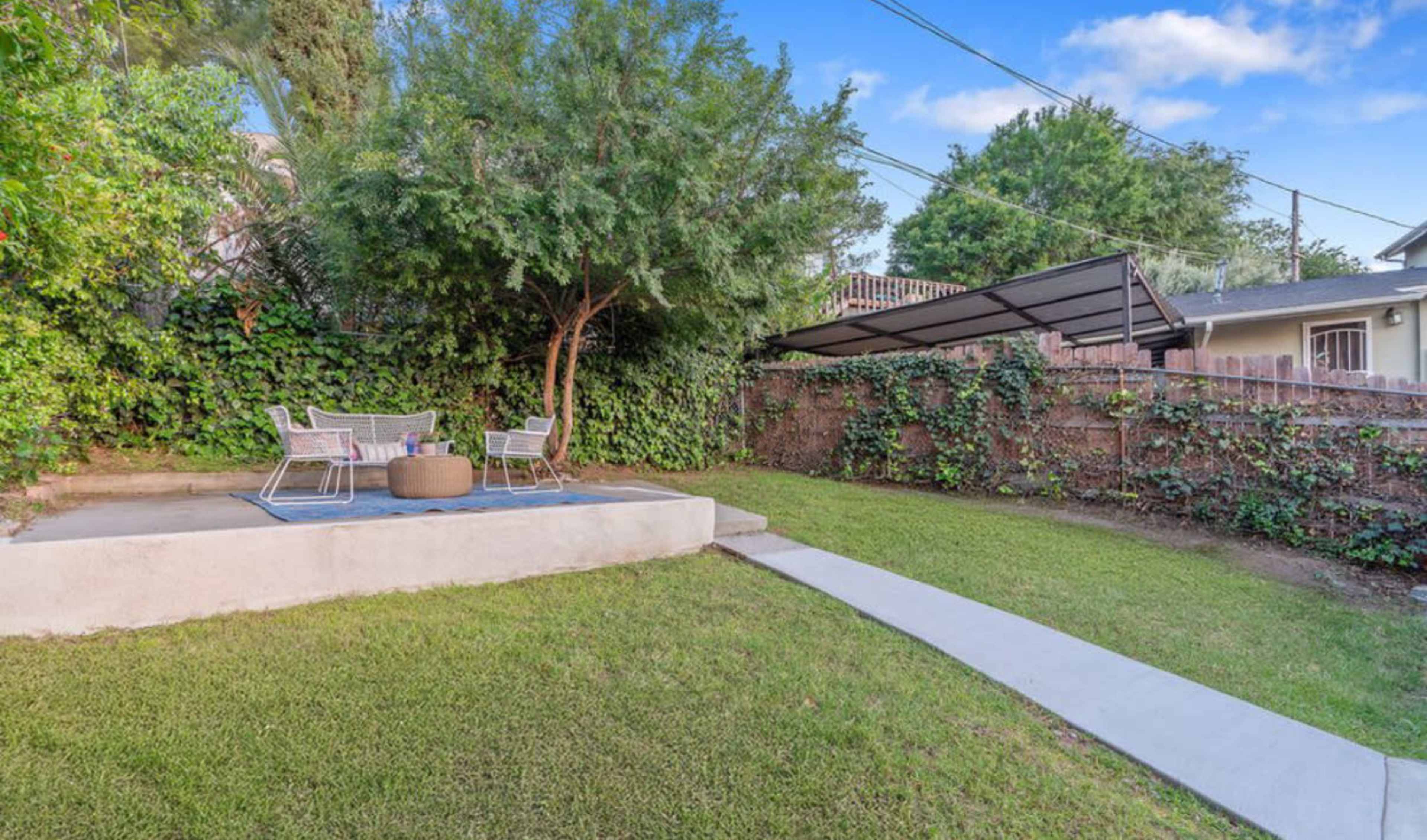 A small outdoor area features two chairs and a table on a concrete patio surrounded by greenery and a neatly trimmed lawn.