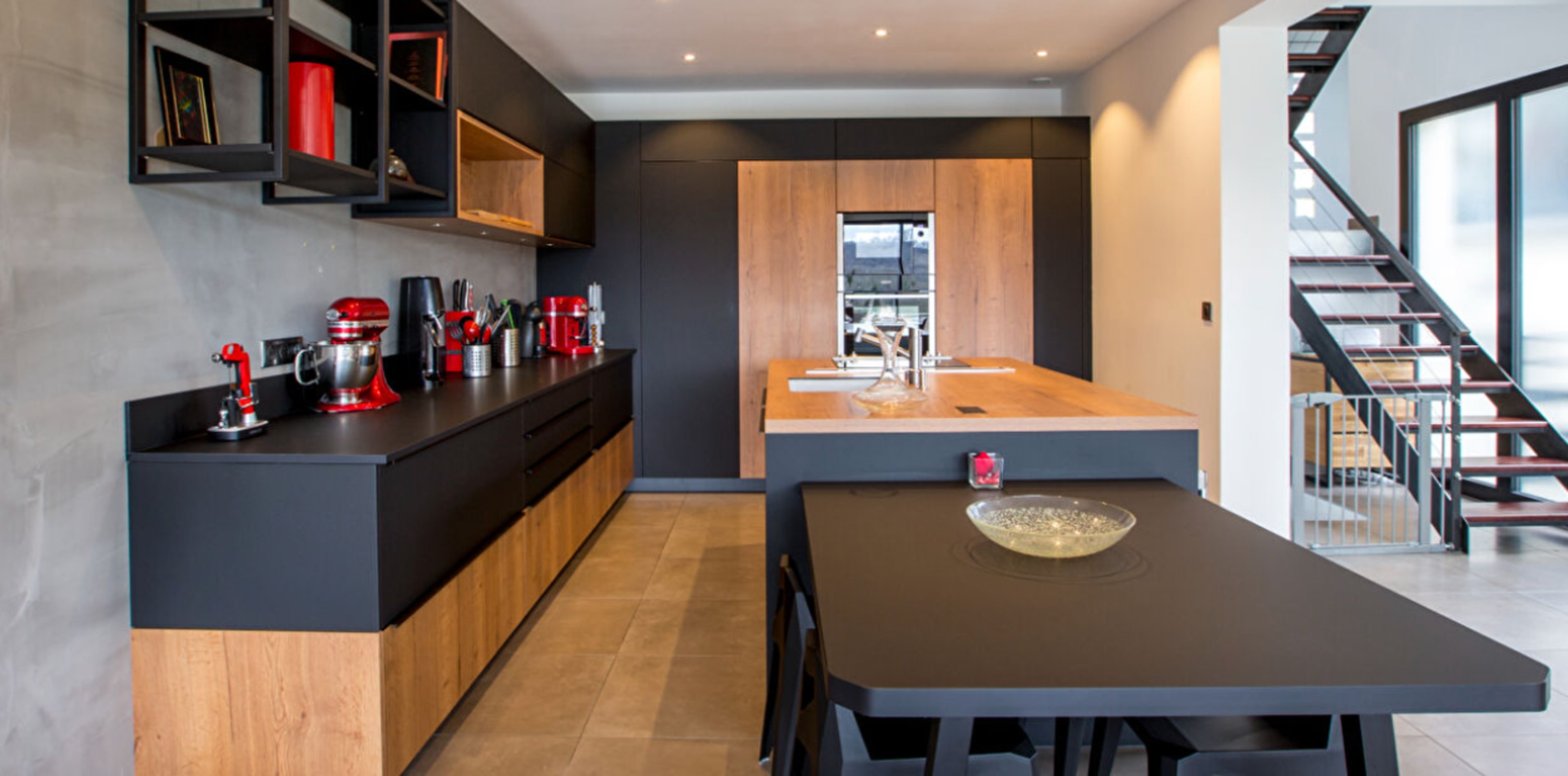 The image shows a modern kitchen with black and wooden cabinetry, featuring a central island and red kitchen appliances.