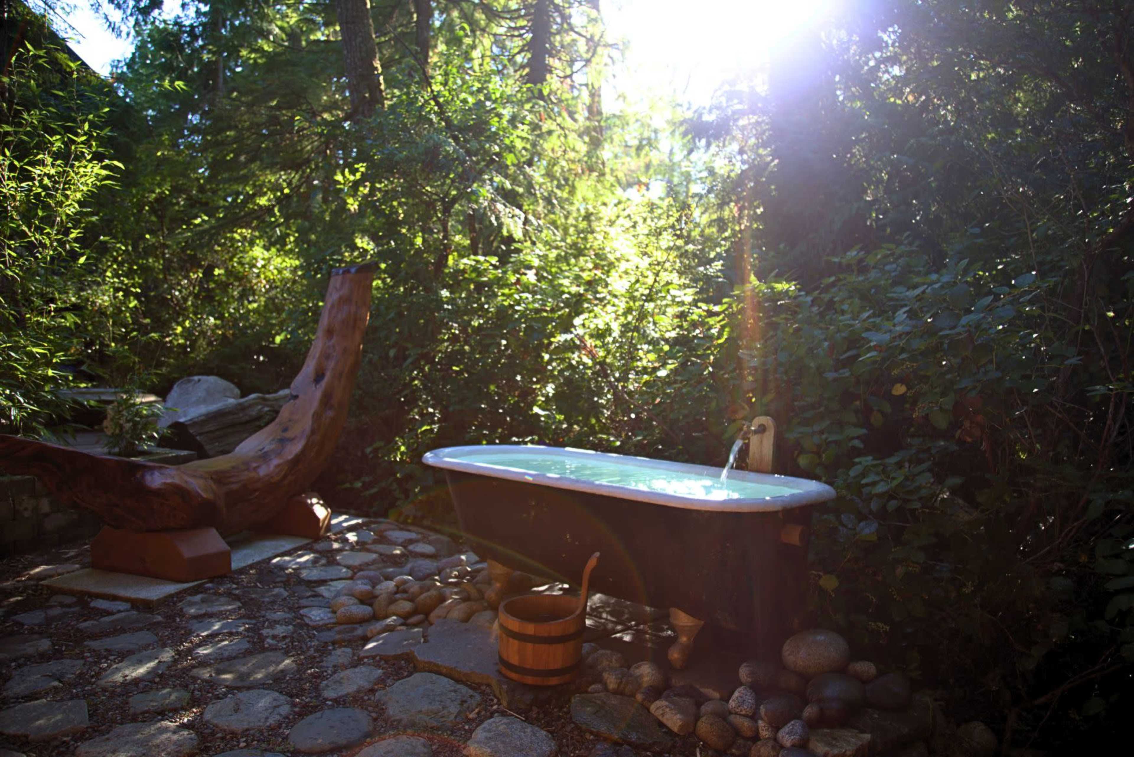 A copper bathtub sits on a stone patio surrounded by greenery, with a wooden chair nearby and sunlight filtering through the trees.