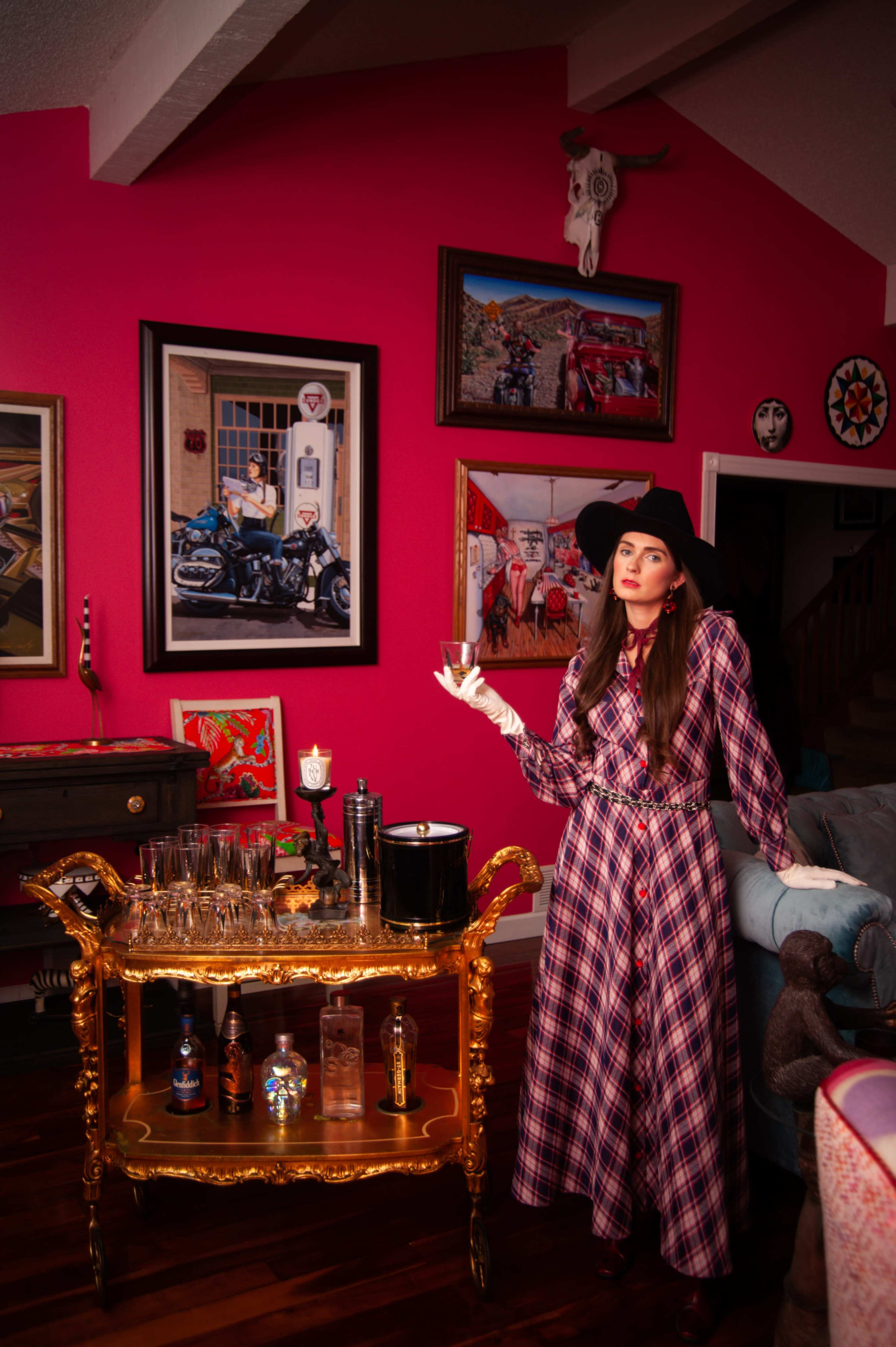 A woman in a plaid dress stands beside a gold bar cart with various bottles and glassware, set against a bright pink wall adorned with framed artwork.