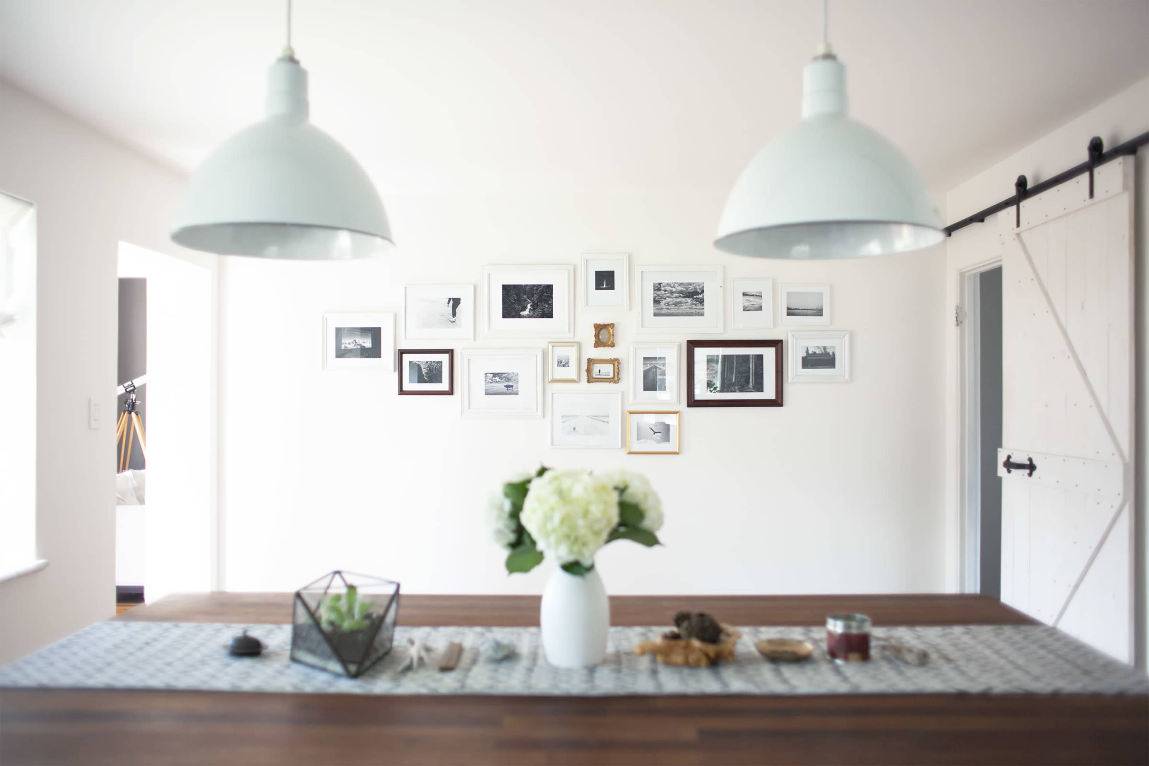 A modern dining area features a wooden table adorned with a floral centerpiece and a patterned runner, while a gallery wall of framed photographs hangs above.