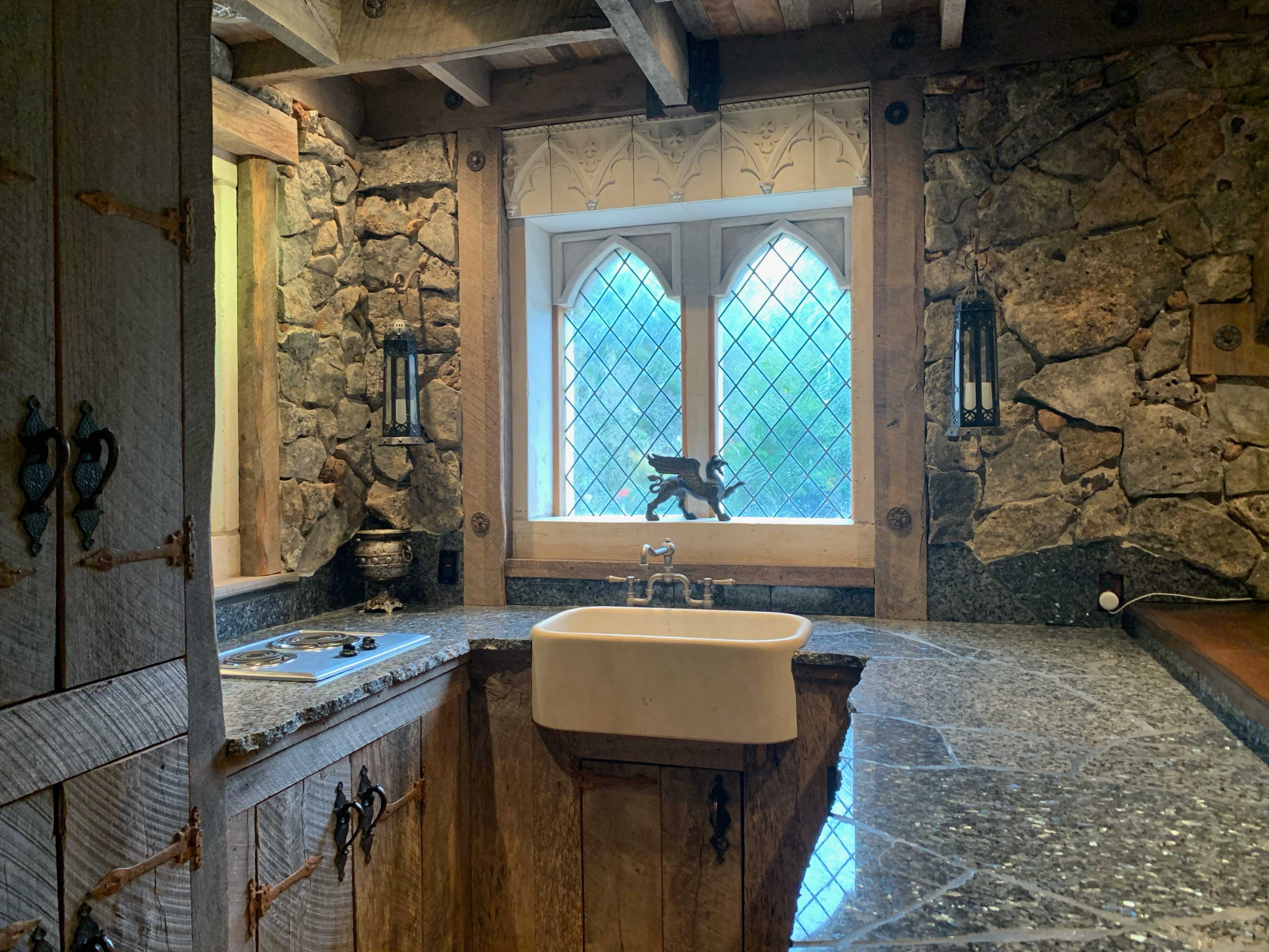 A rustic kitchen featuring a stone wall, a deep sink, and a large window with a decorative frame.