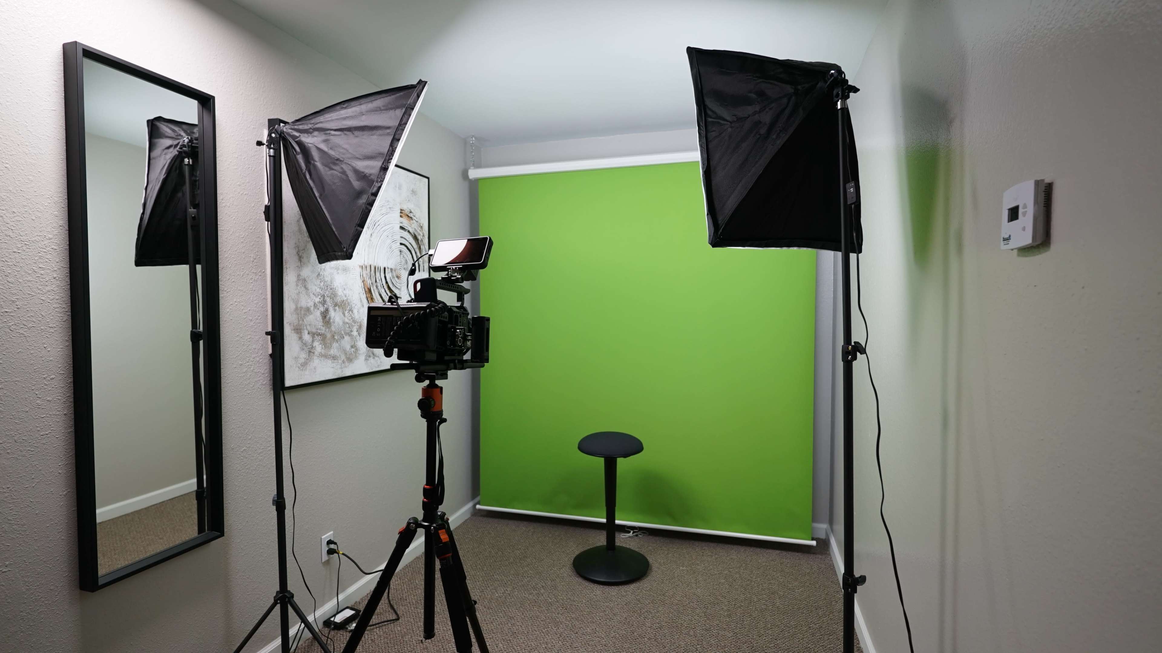 A small studio setup with a camera on a tripod, two softbox lights, a green backdrop, and a black stool.