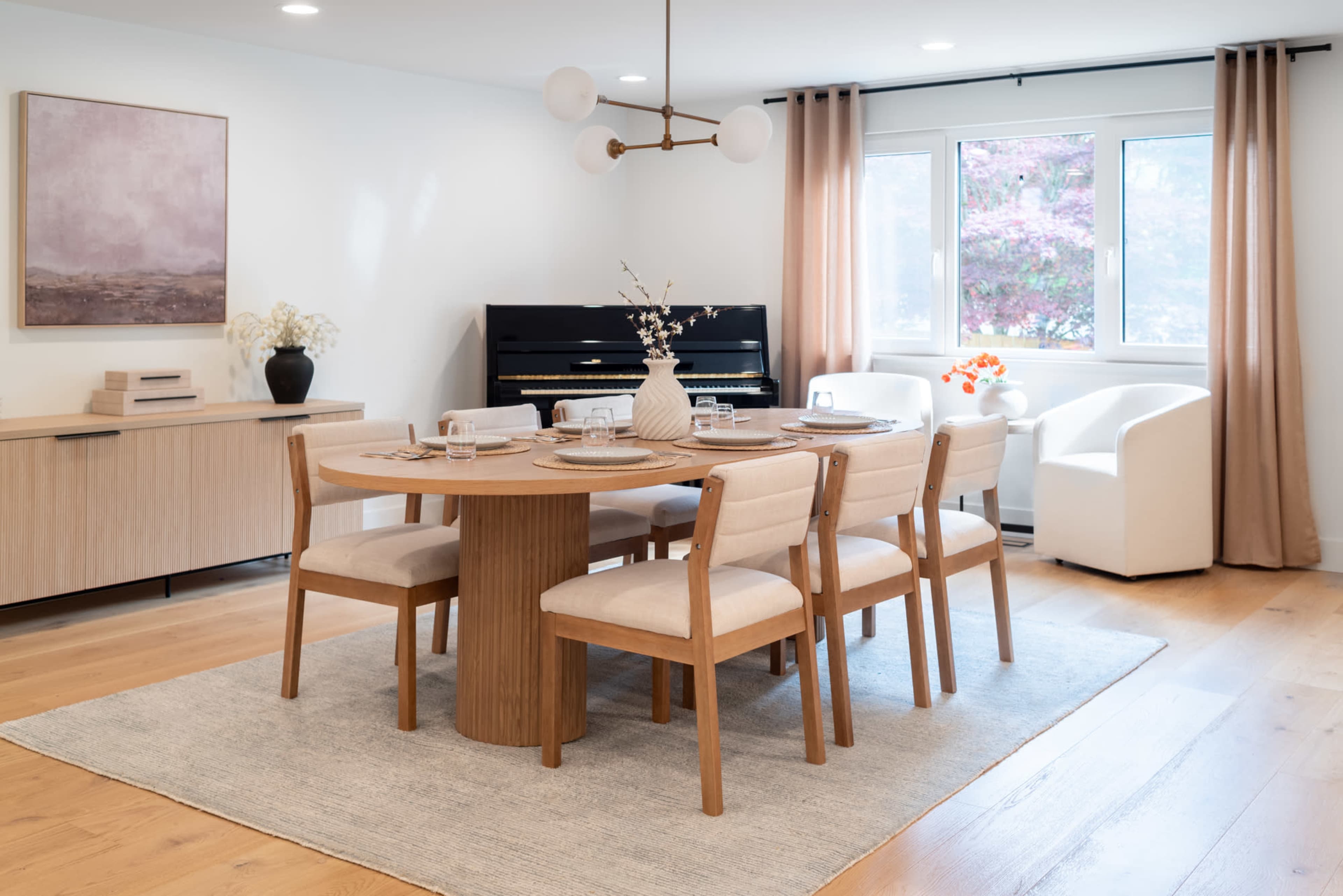 A modern dining area features a circular wooden table with six chairs, a piano in the corner, and large windows covered by light curtains.