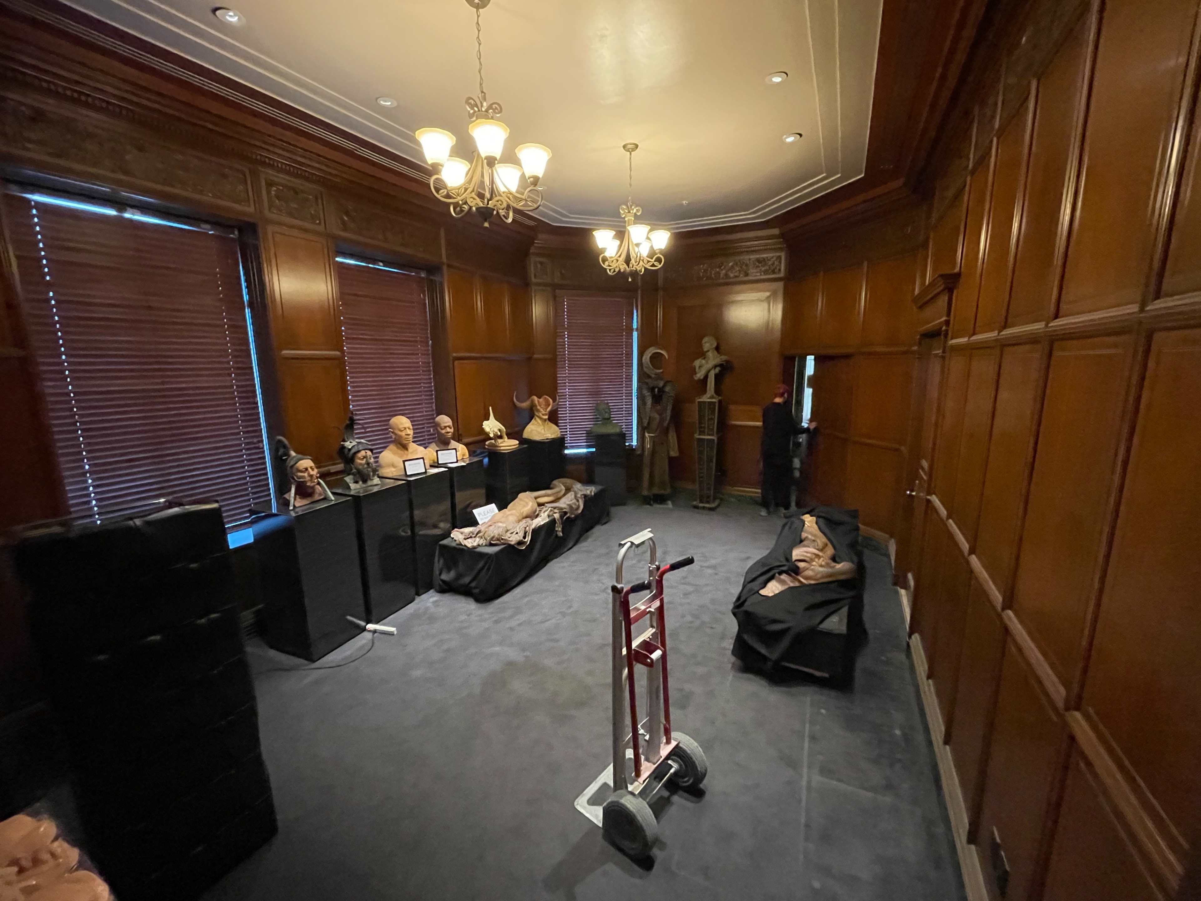 The room features wooden paneling, a chandelier, and various sculptures displayed on tables, with a doll or mannequin lying on a black cloth in the center.