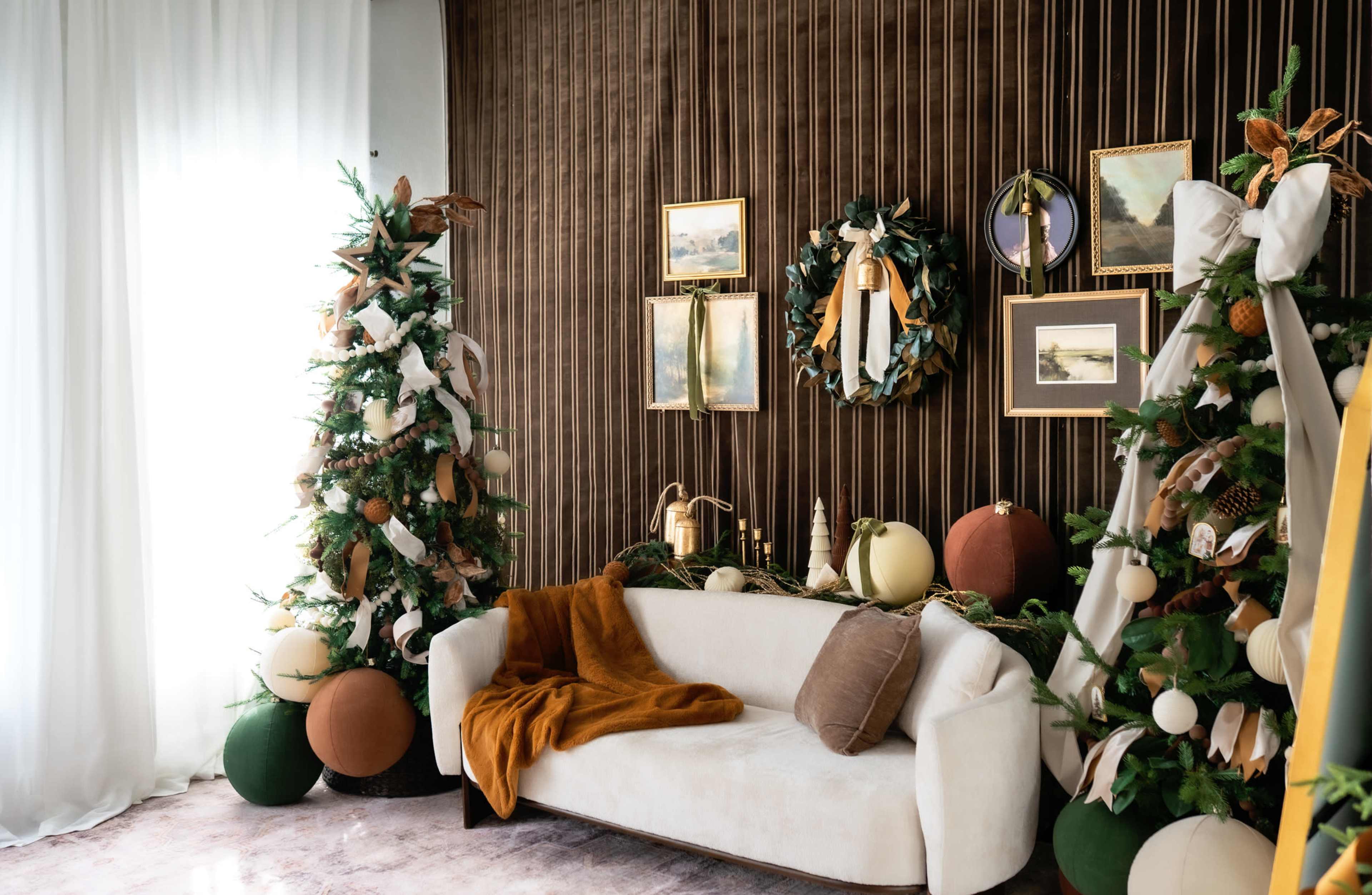 A decorated living room features two Christmas trees flanking a white sofa, with wall art and garlands hanging above.