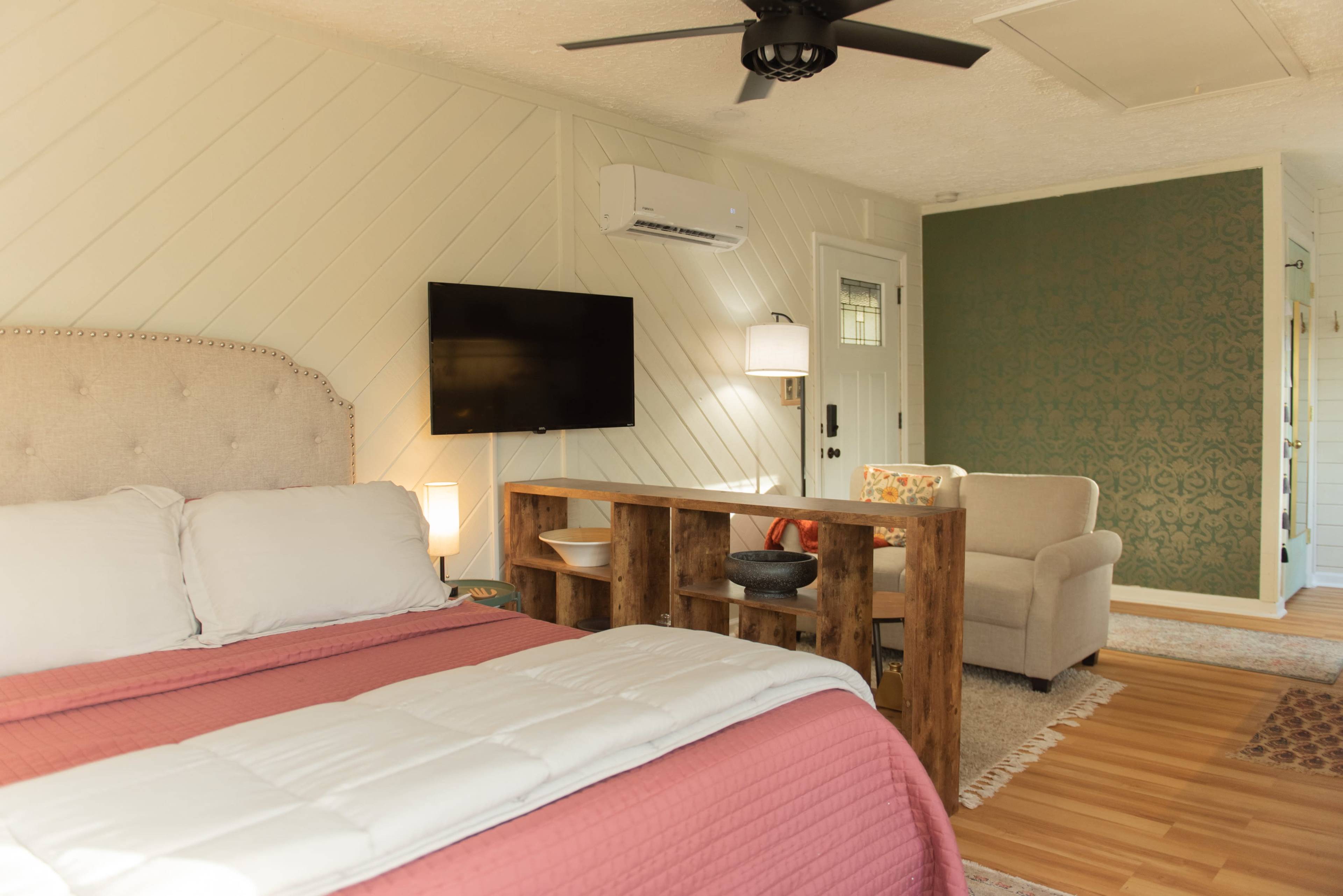A cozy bedroom featuring a queen-sized bed with a pink quilt, a wooden partition with decorative items, a flat-screen TV mounted on the wall, and a chair in the corner.