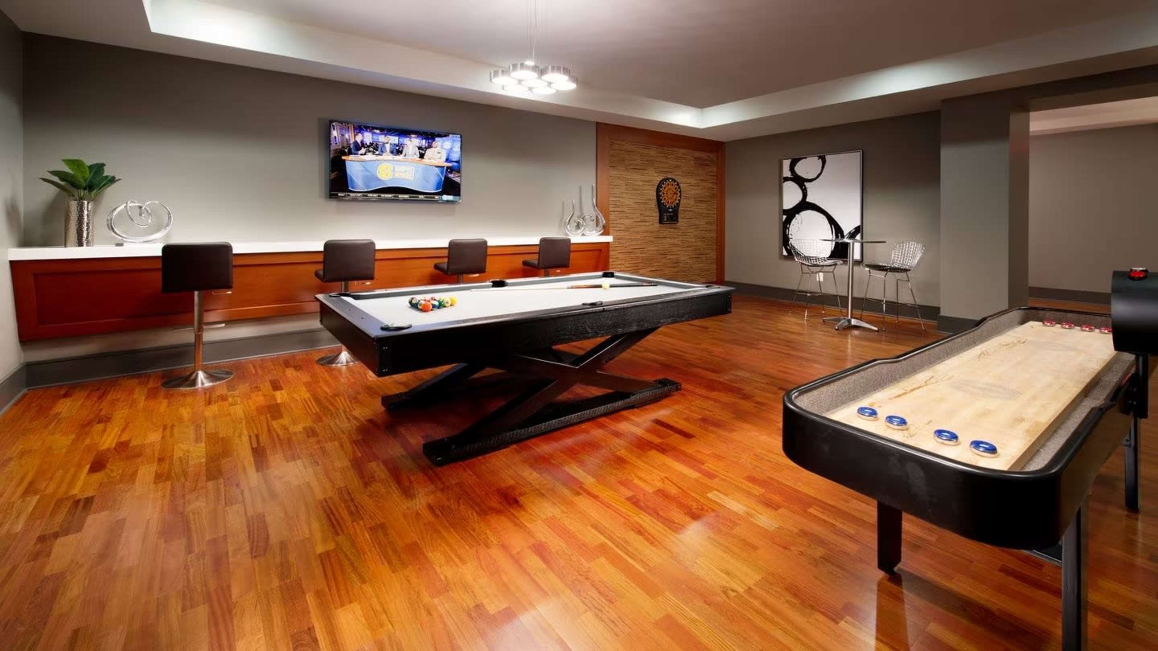 A recreational room with a pool table, a shuffleboard table, bar stools, and a television mounted on the wall.