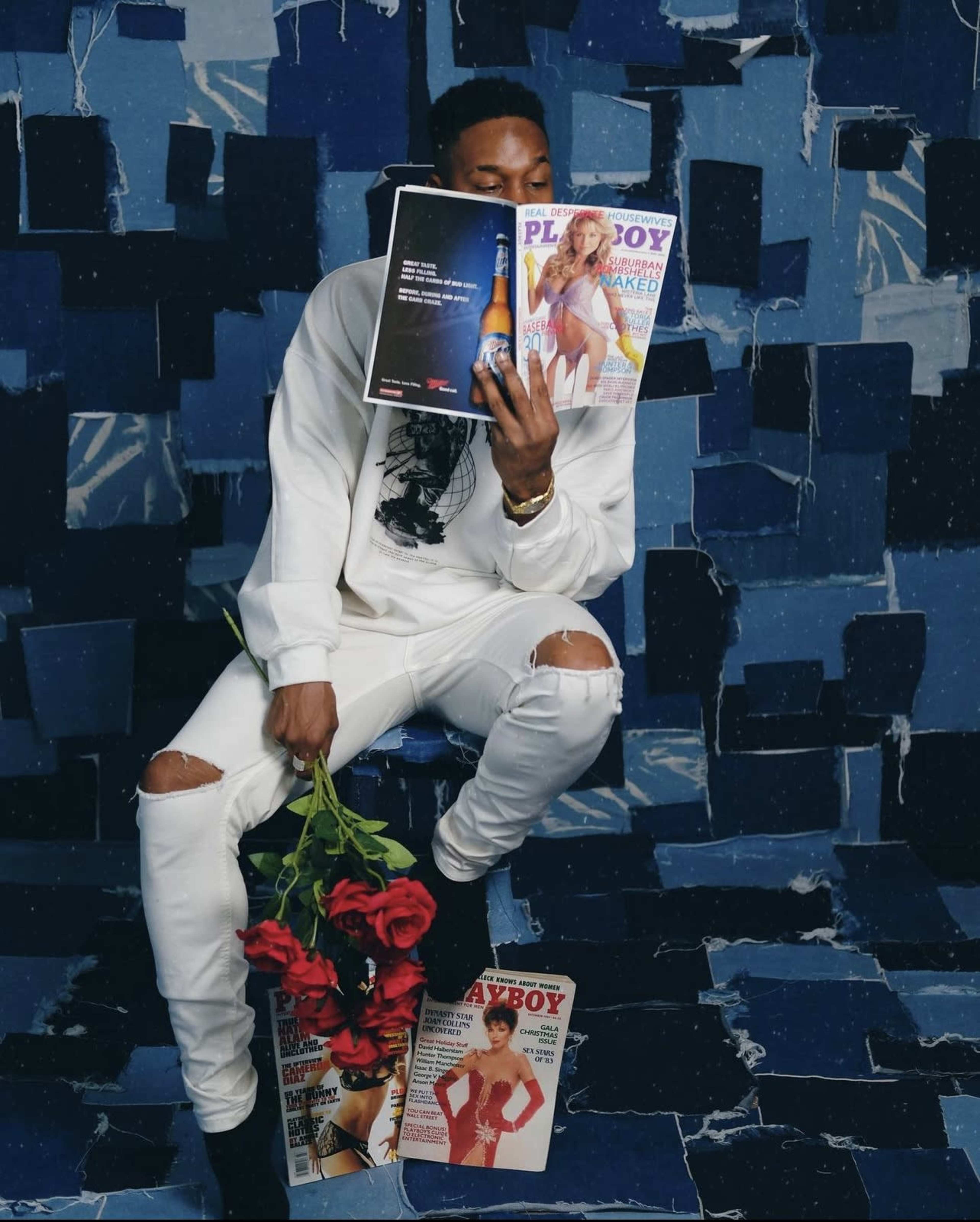 A person in white clothing sits on a collage of blue paper, holding a magazine in front of their face and a bouquet of roses in their other hand, with additional magazines laid on the ground.