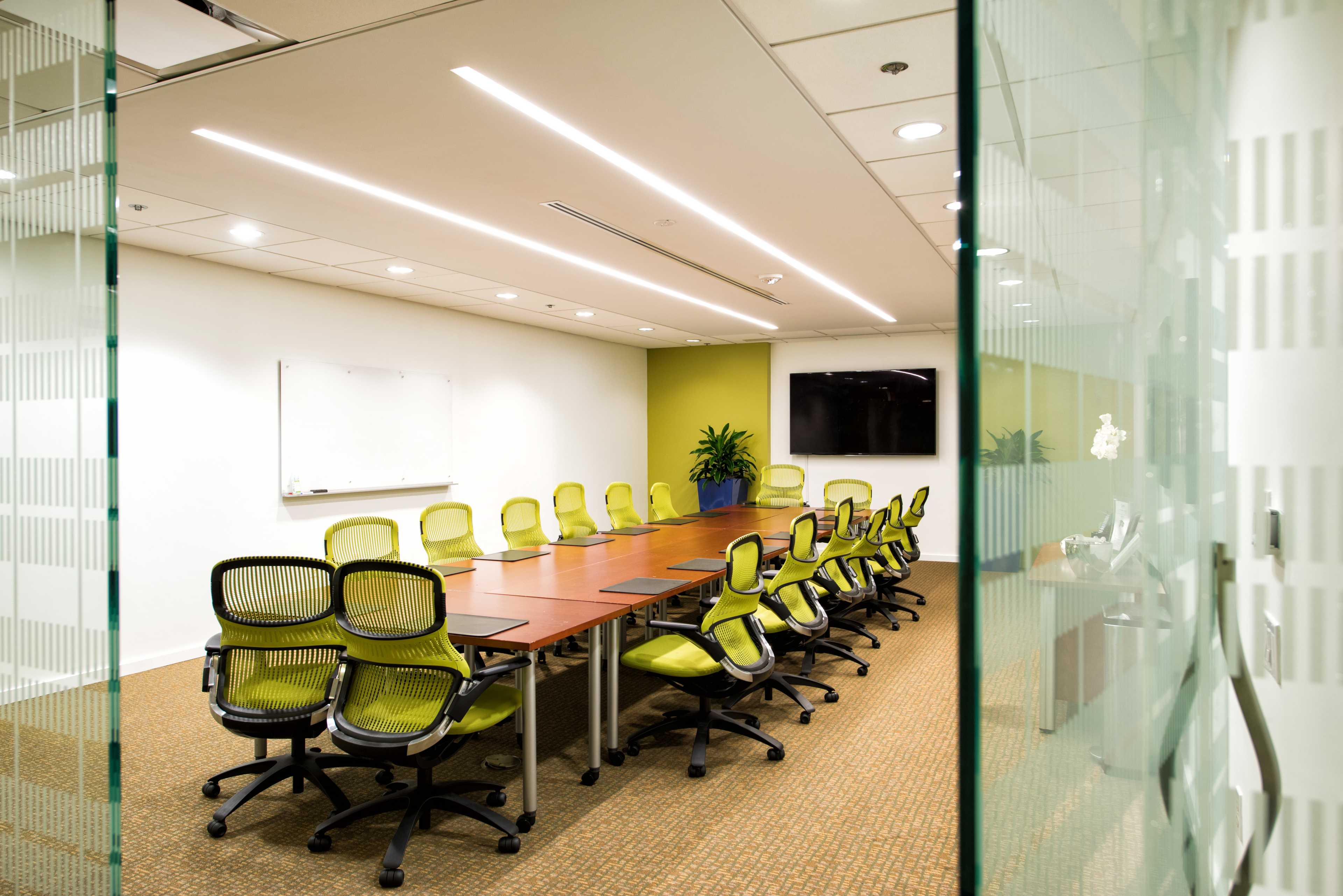 A large conference room features a long wooden table surrounded by green ergonomic chairs, with a whiteboard and a TV on the wall.