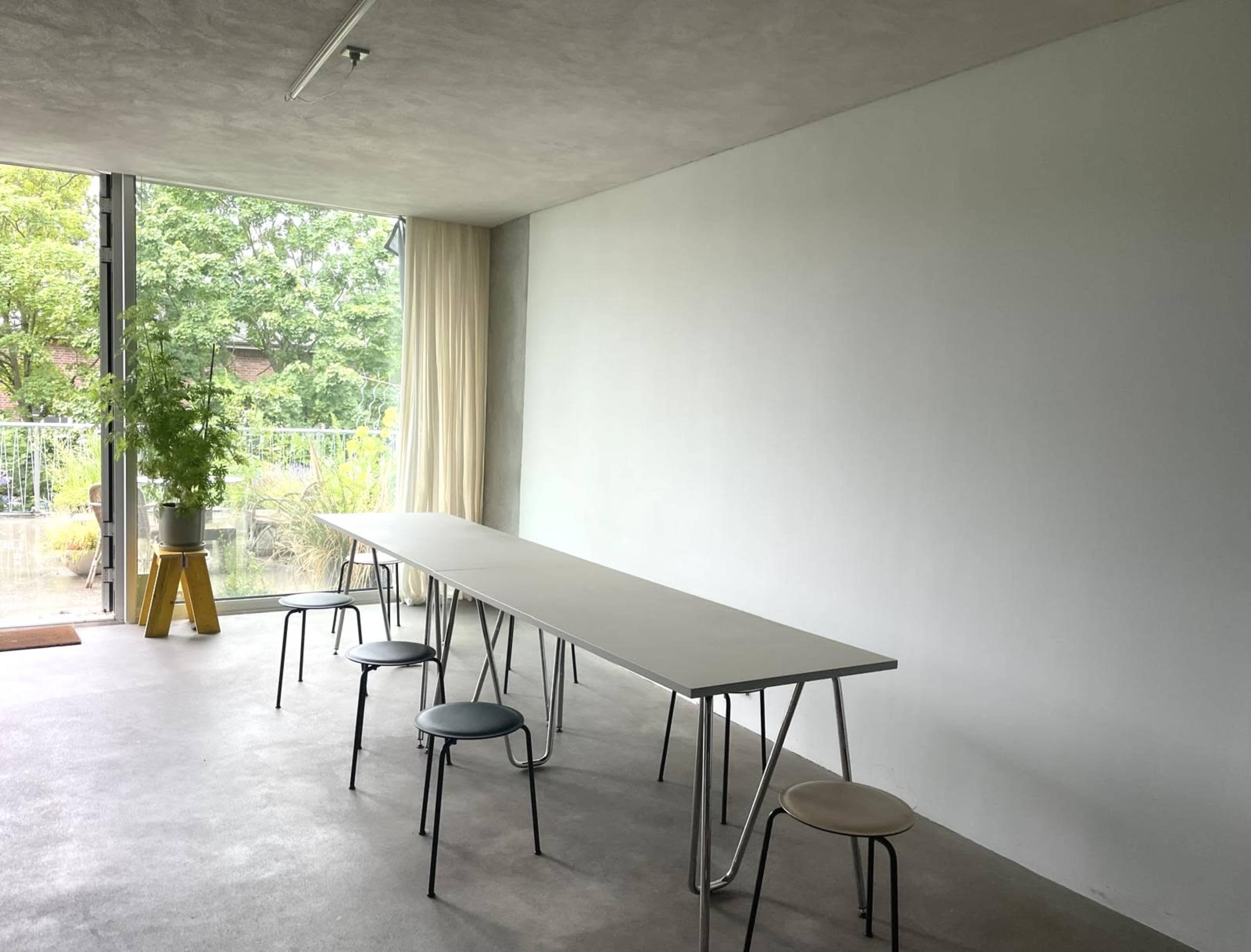 The image shows a minimalist room with a long table and several chairs, facing a large window that overlooks greenery.