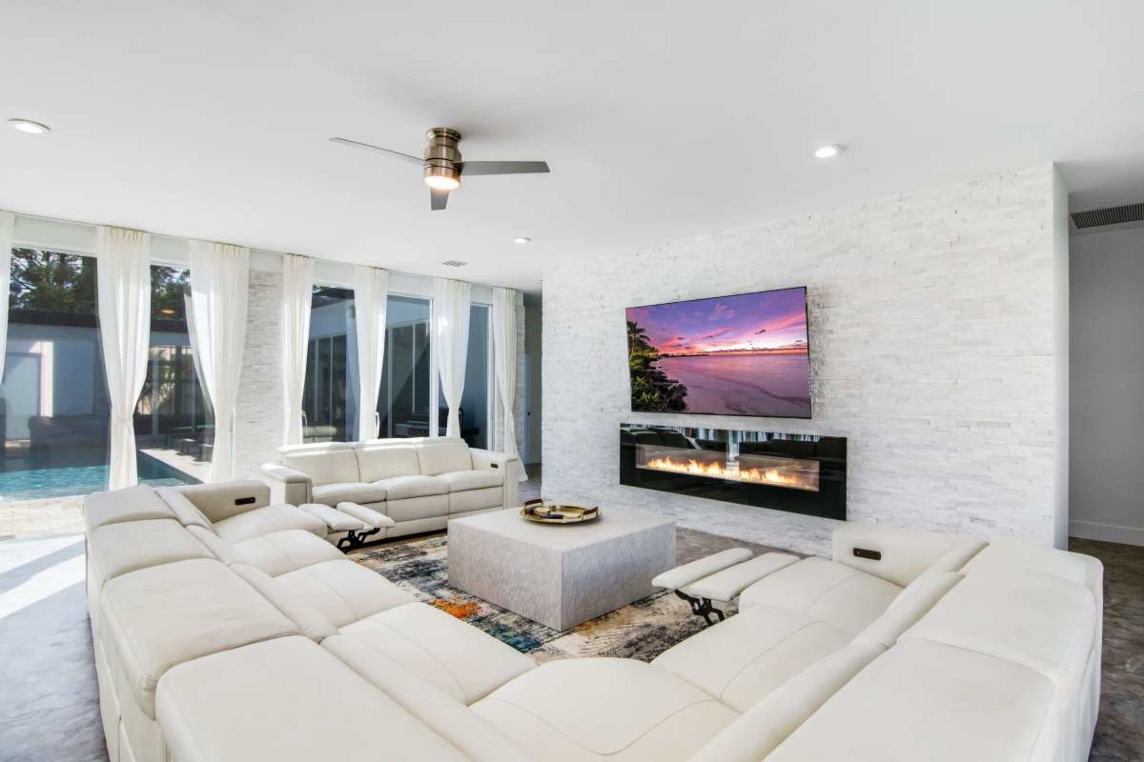A modern living room features a sectional sofa, a coffee table, a wall-mounted television, and a fireplace, with large windows overlooking a pool area.