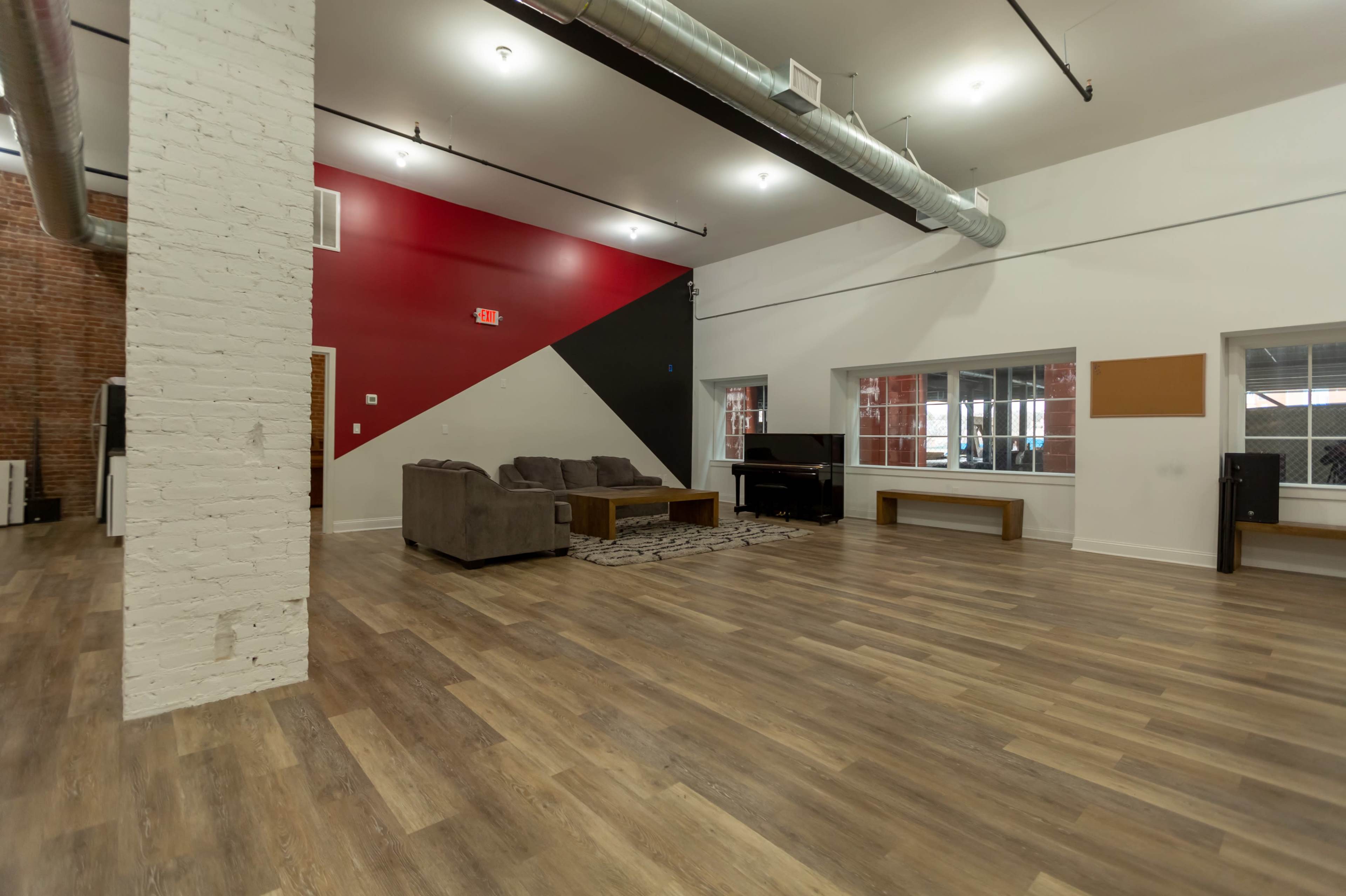 The room features a spacious layout with wooden flooring, a red and black accent wall, and furnishings including a gray couch and a piano.