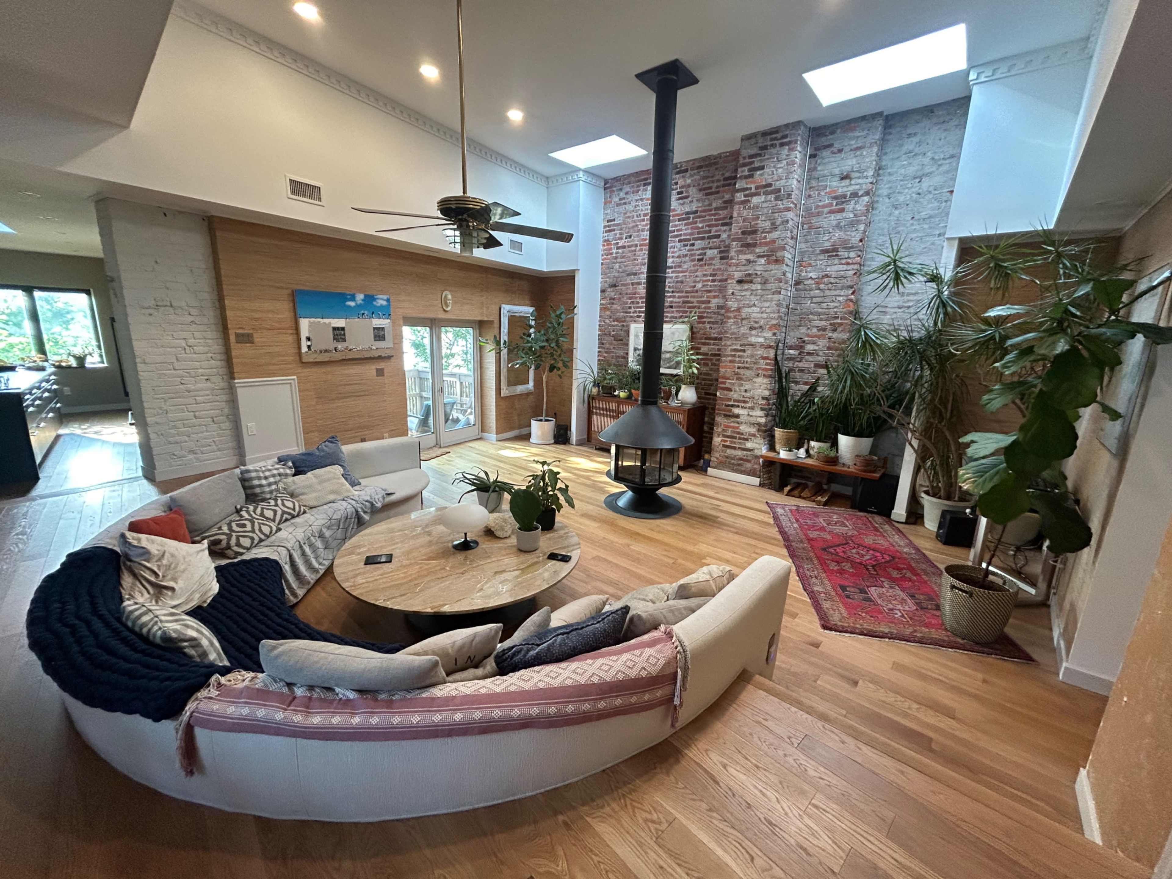 A modern living room features a circular white couch, a central round table, a metal wood stove, and large windows with exposed brick walls and indoor plants.