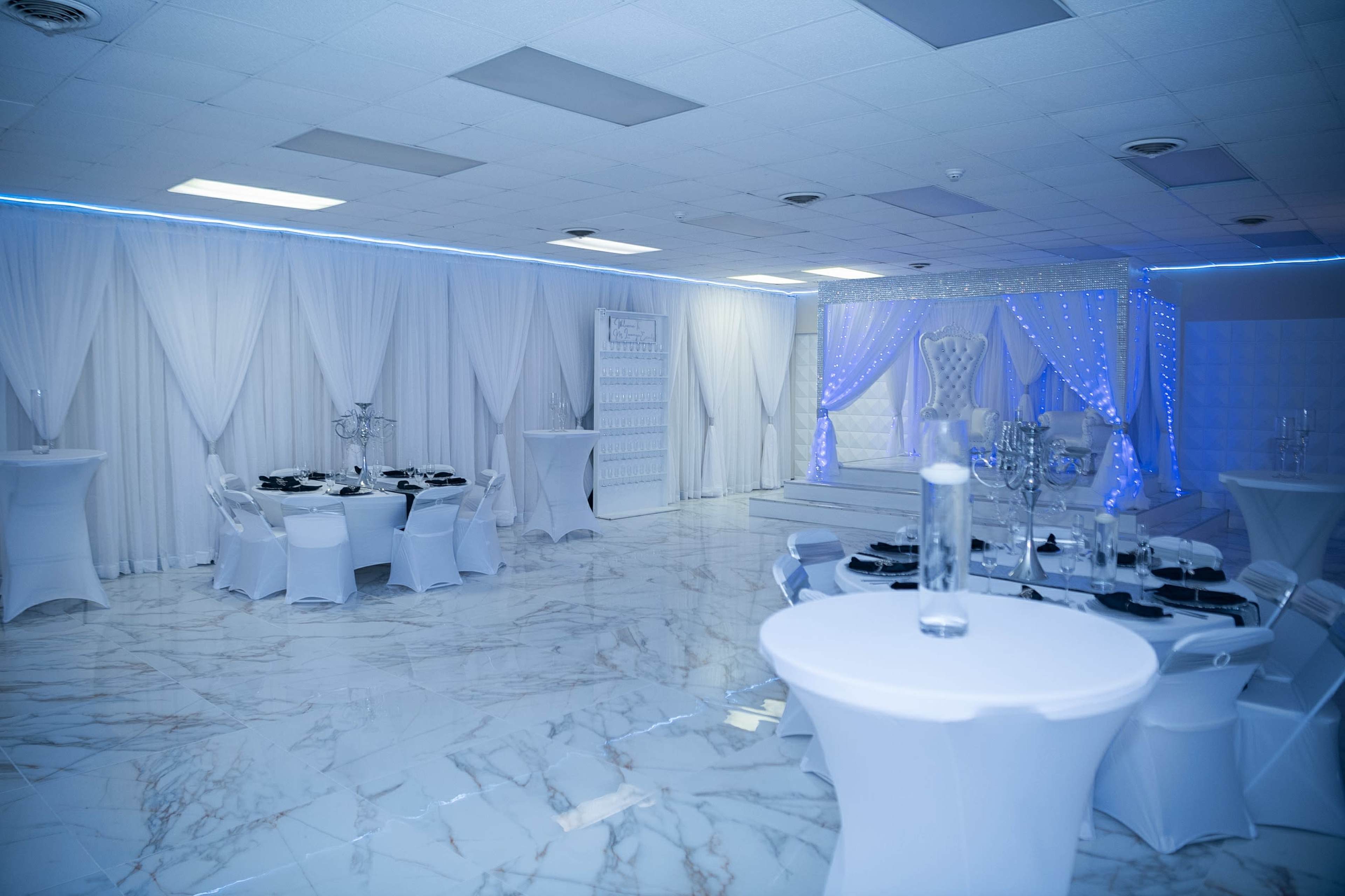 A spacious banquet hall with white drapes, marble flooring, and elegantly arranged tables set for an event.
