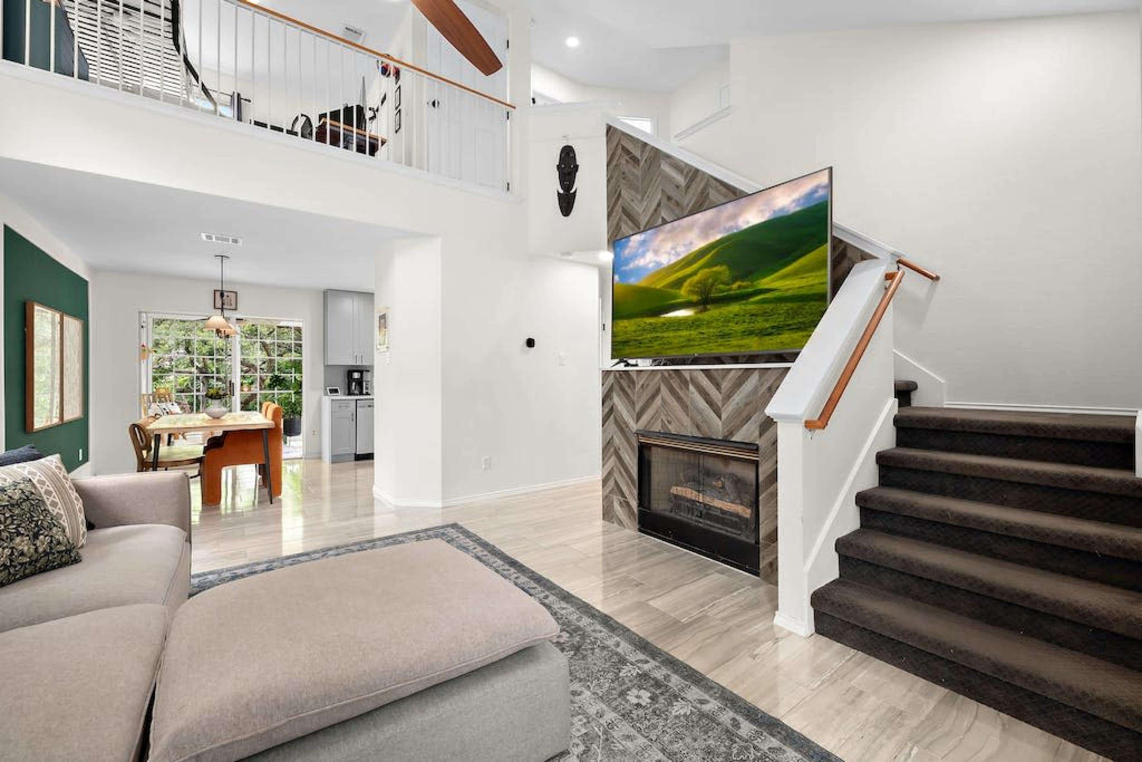 A modern living room features a sectional sofa, a large television mounted on a textured wall, and a staircase leading to the upper level.