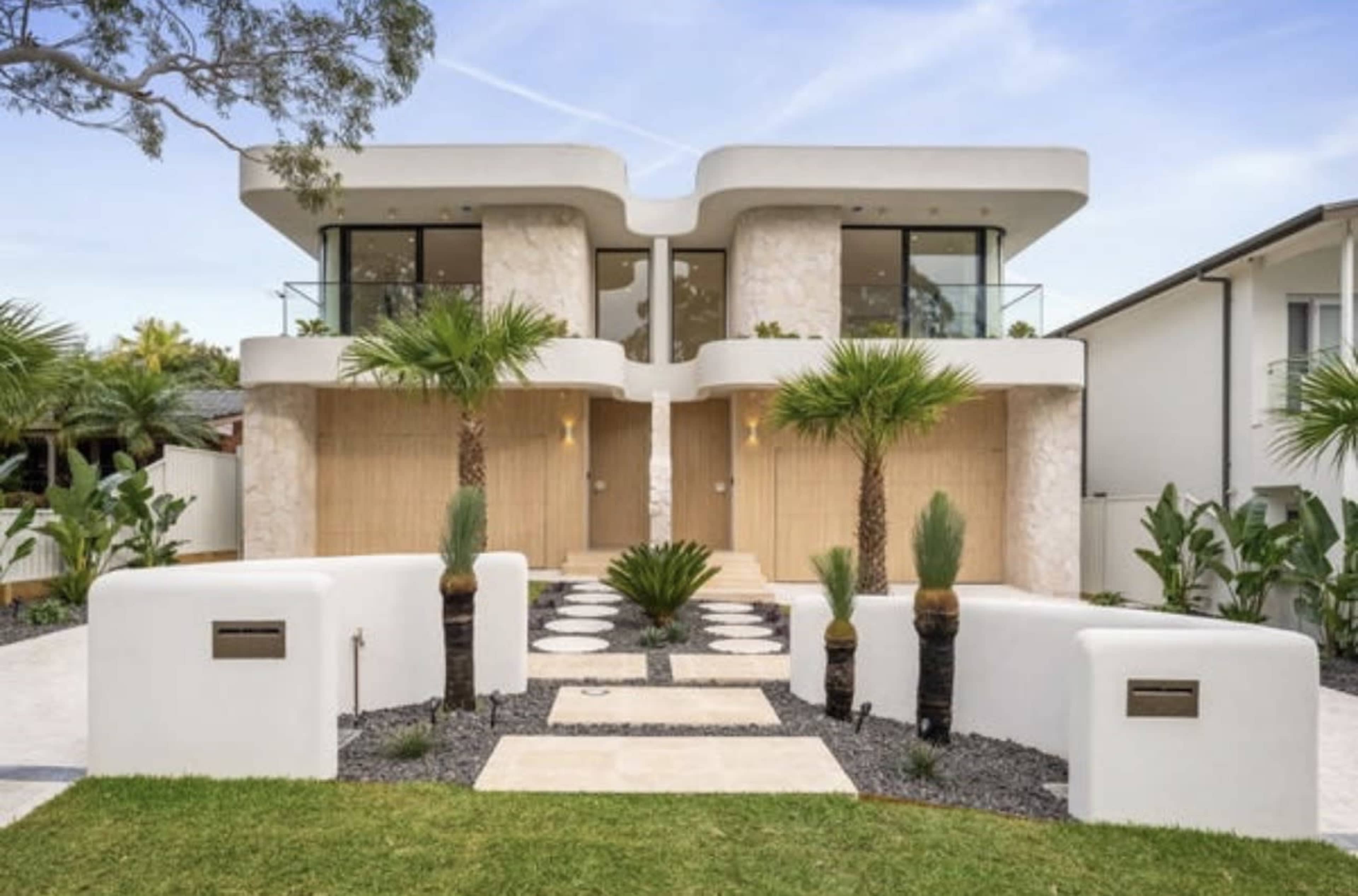 The image features a contemporary two-story house with a symmetrical façade, large glass windows, and a landscaped front yard with palm trees and stone pathways.