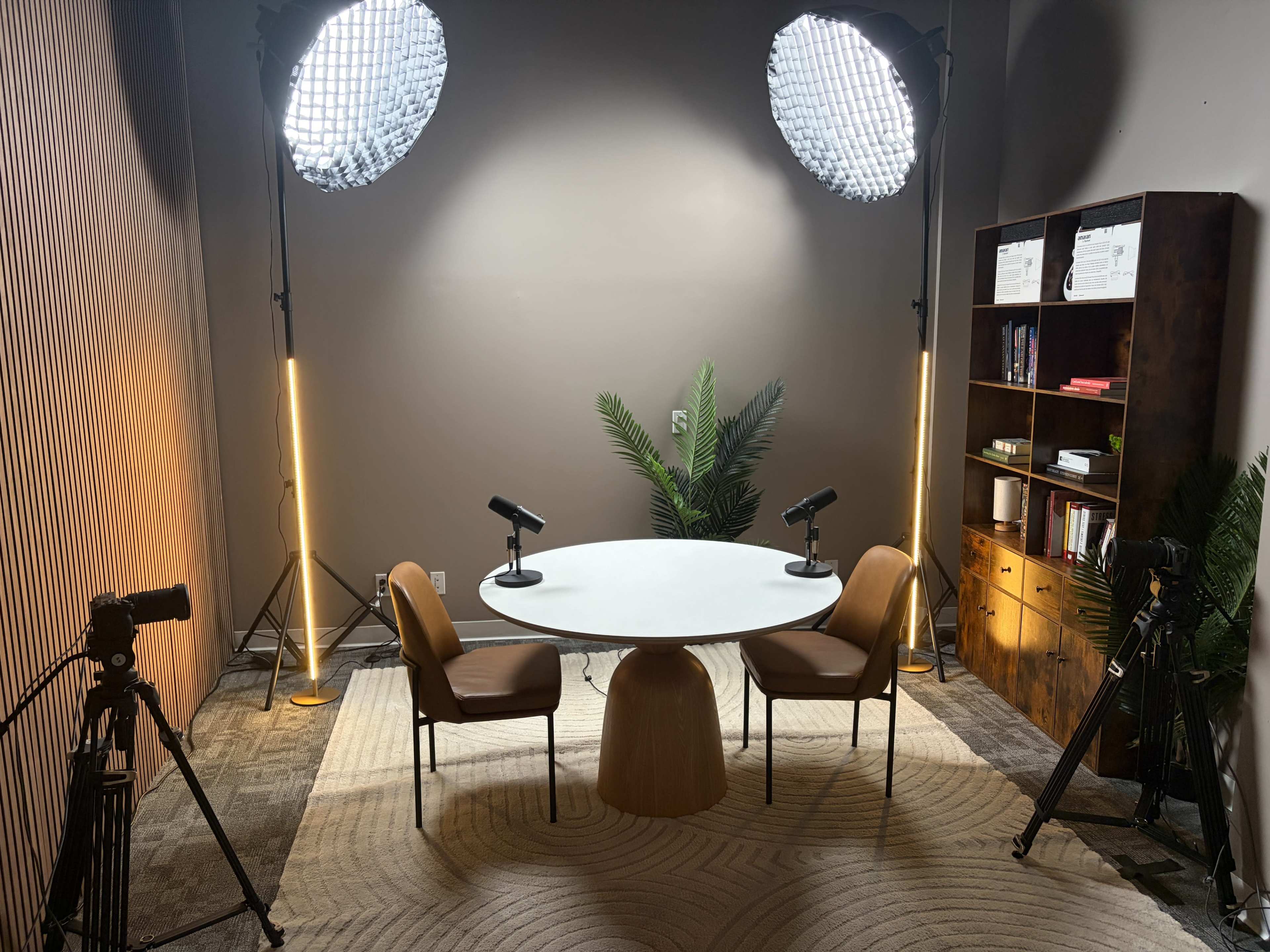 The image shows a podcast recording setup featuring a round white table with two chairs, two microphones, and studio lights, surrounded by a bookshelf and decorative plants.