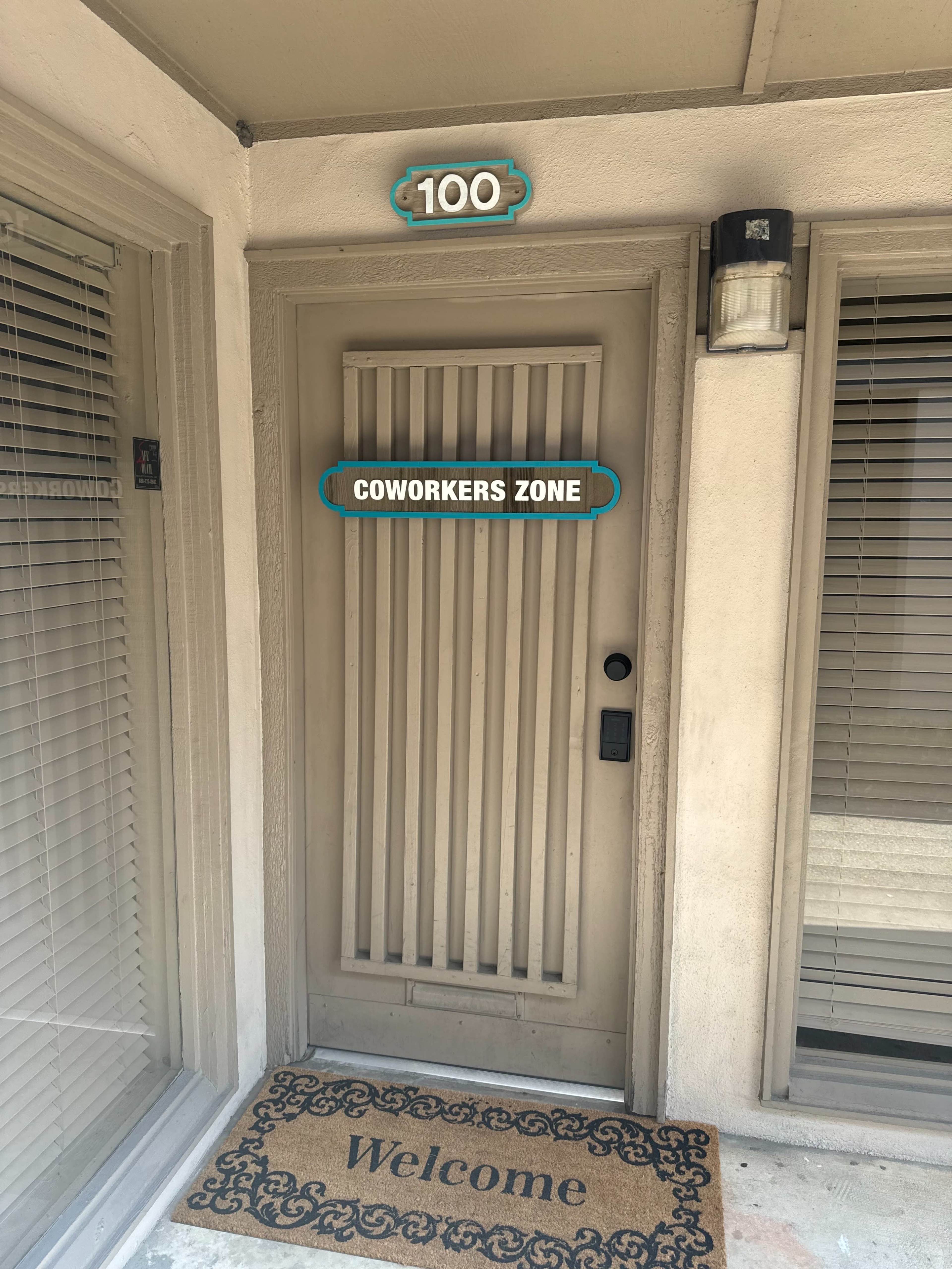 The image shows a beige door with the number "100," a sign that reads "COWORKERS ZONE," and a welcome mat in front.