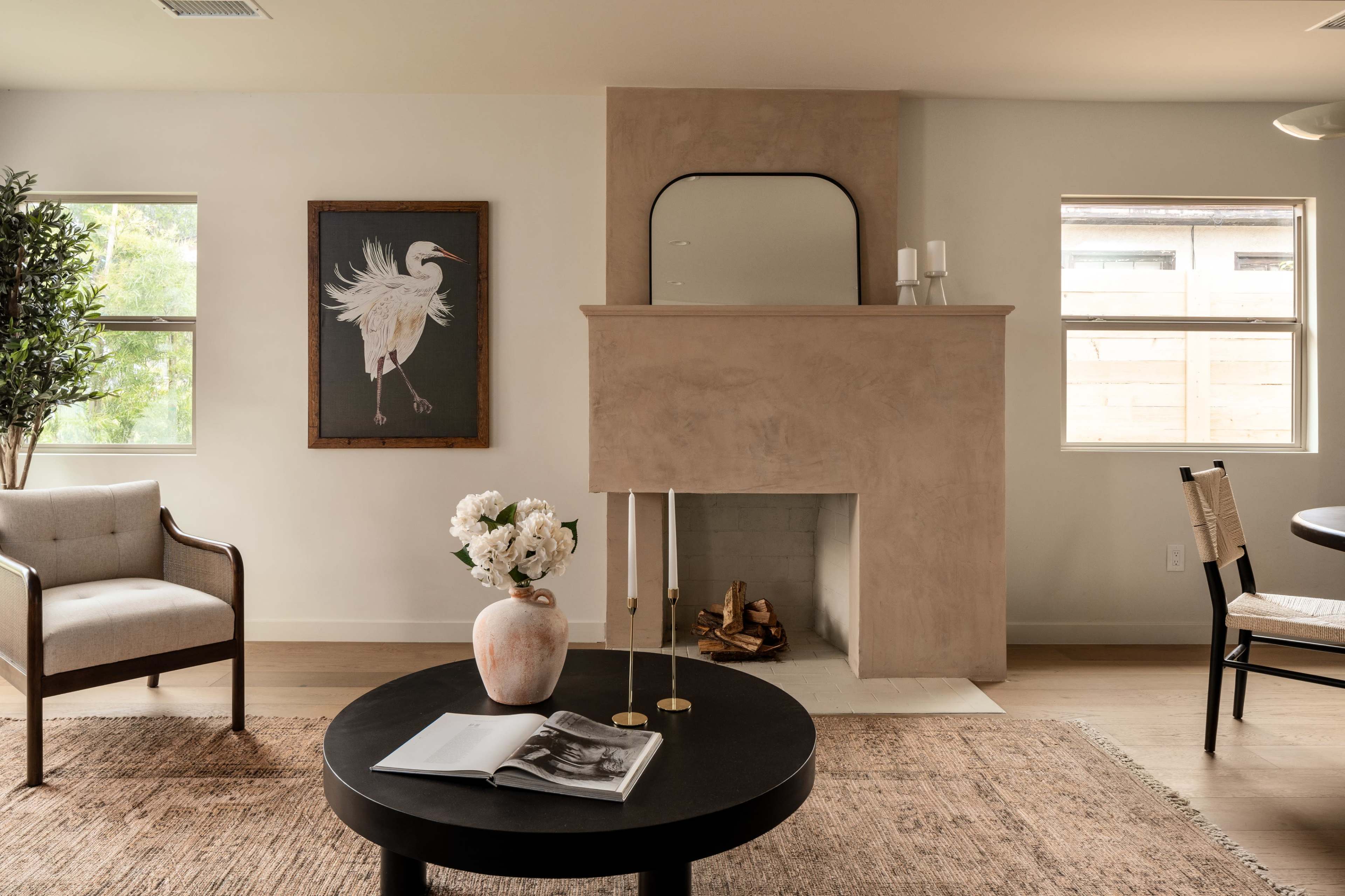 A modern living room with a minimalist design featuring a fireplace, a round coffee table with a book, and a framed artwork of a bird on the wall.
