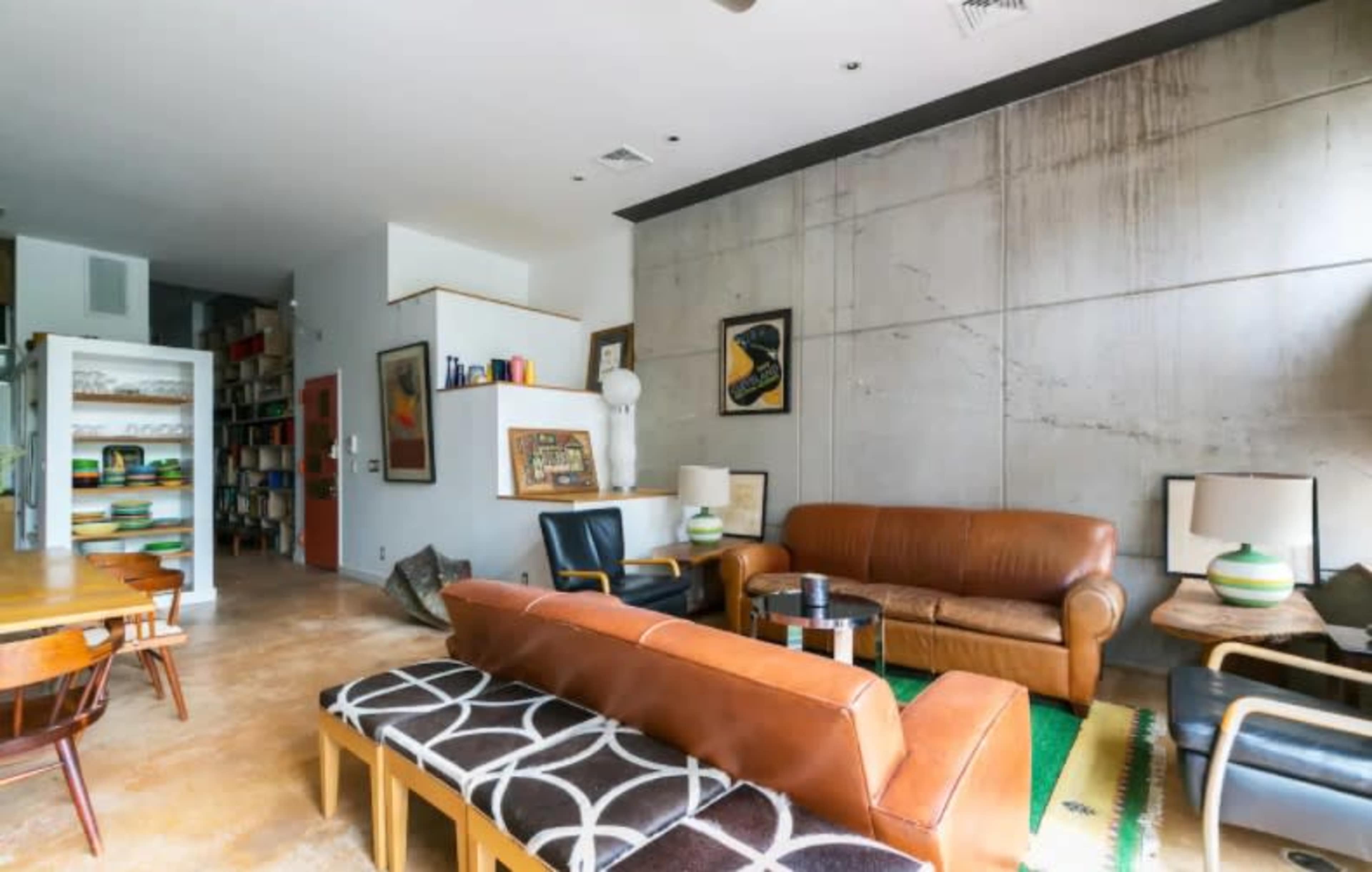 A spacious living area featuring a brown leather sofa, mid-century chairs, a concrete wall, and a dining table with wooden chairs.