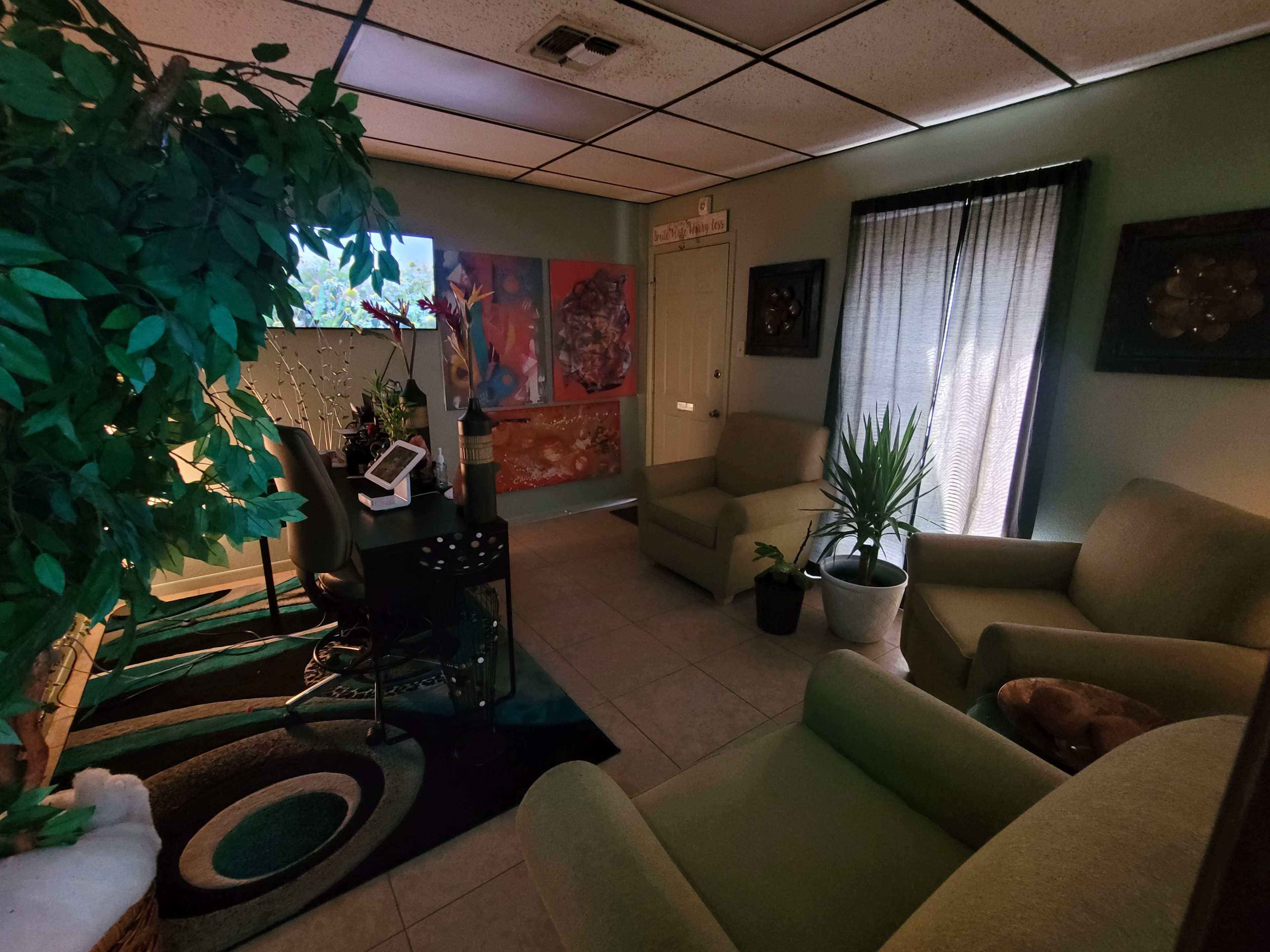 A cozy office space with two green chairs, a desk, artwork on the walls, and a large plant in a corner.