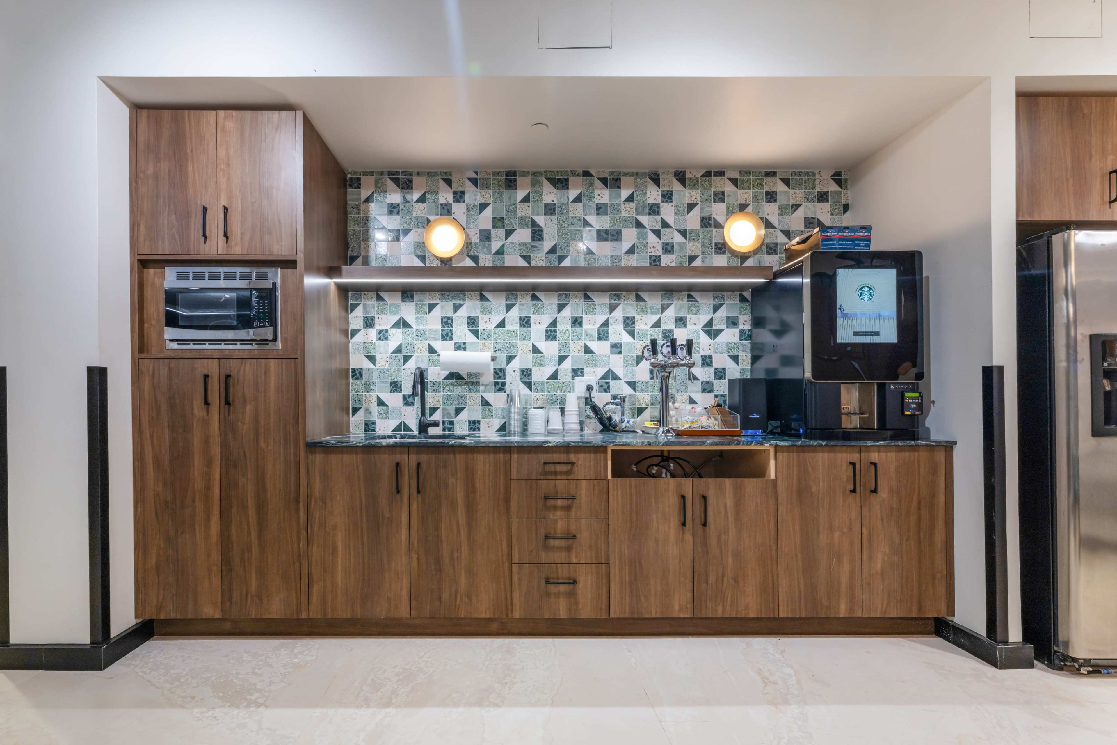 The image shows a modern kitchen area with wooden cabinets, a microwave, a coffee machine, and a decorative tiled backsplash.