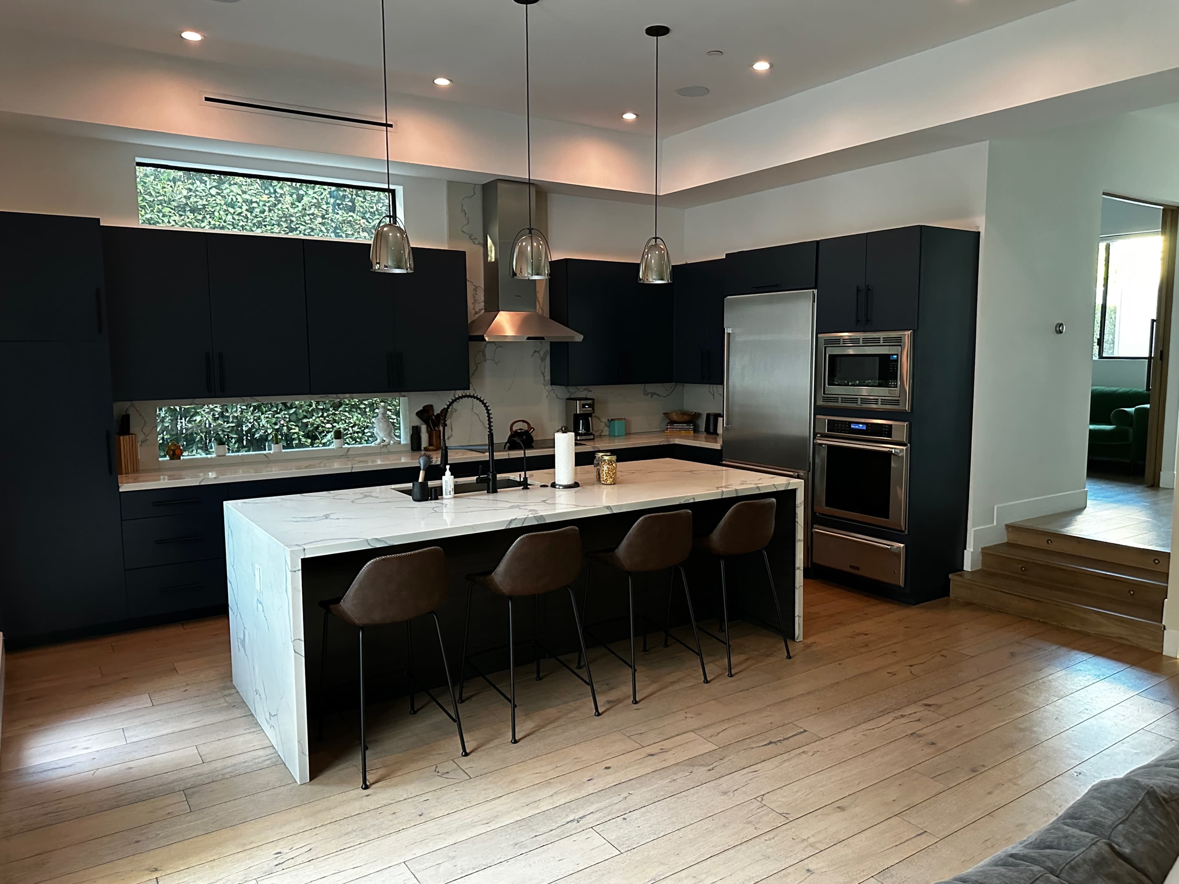 A modern kitchen with dark cabinetry, a large marble island with seating, and stainless steel appliances, illuminated by pendant lights.
