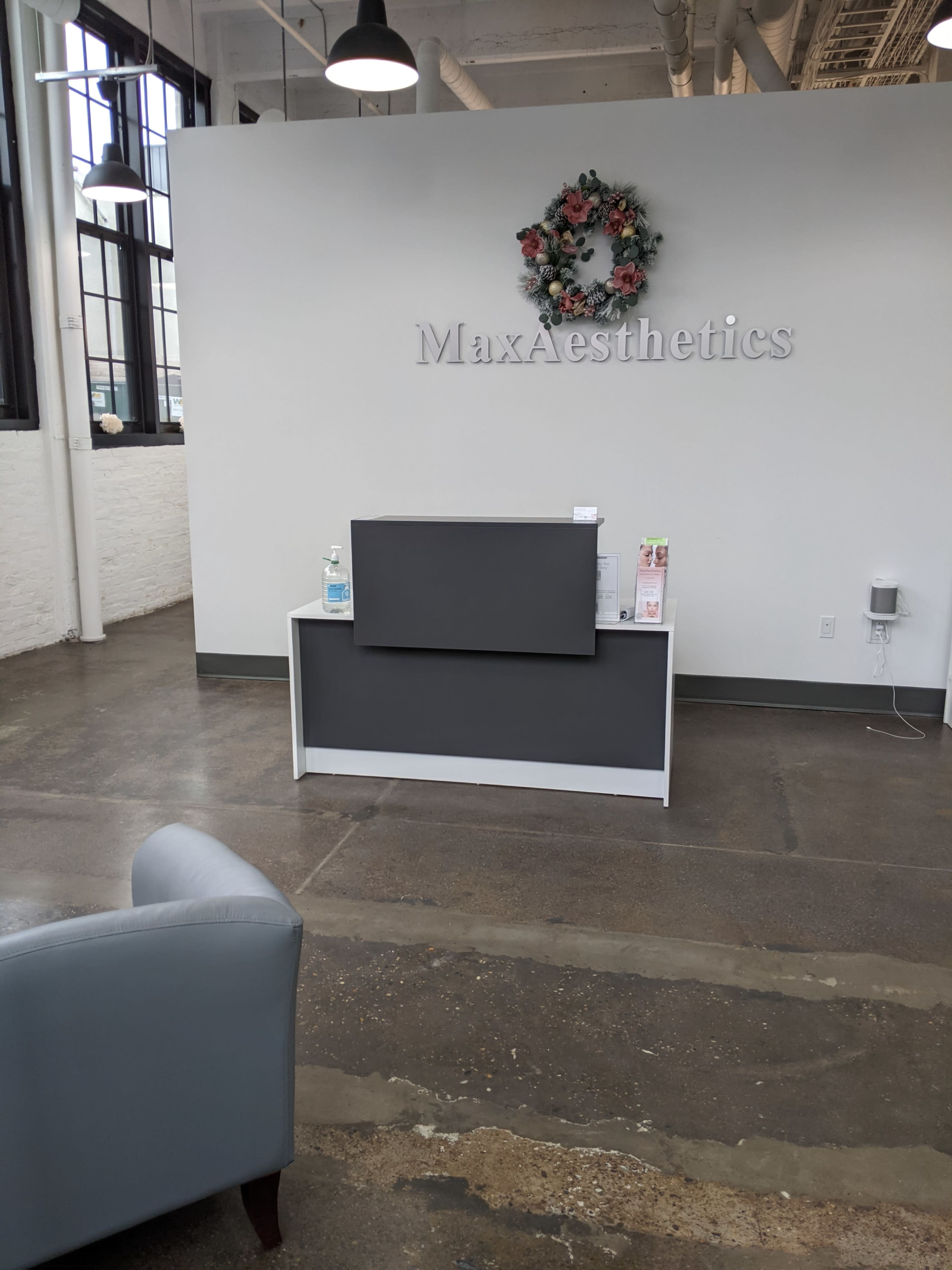 The image shows a reception area of a business named MaxAesthetics, featuring a grey reception desk, a decorative floral wreath on the wall, and large windows providing natural light.