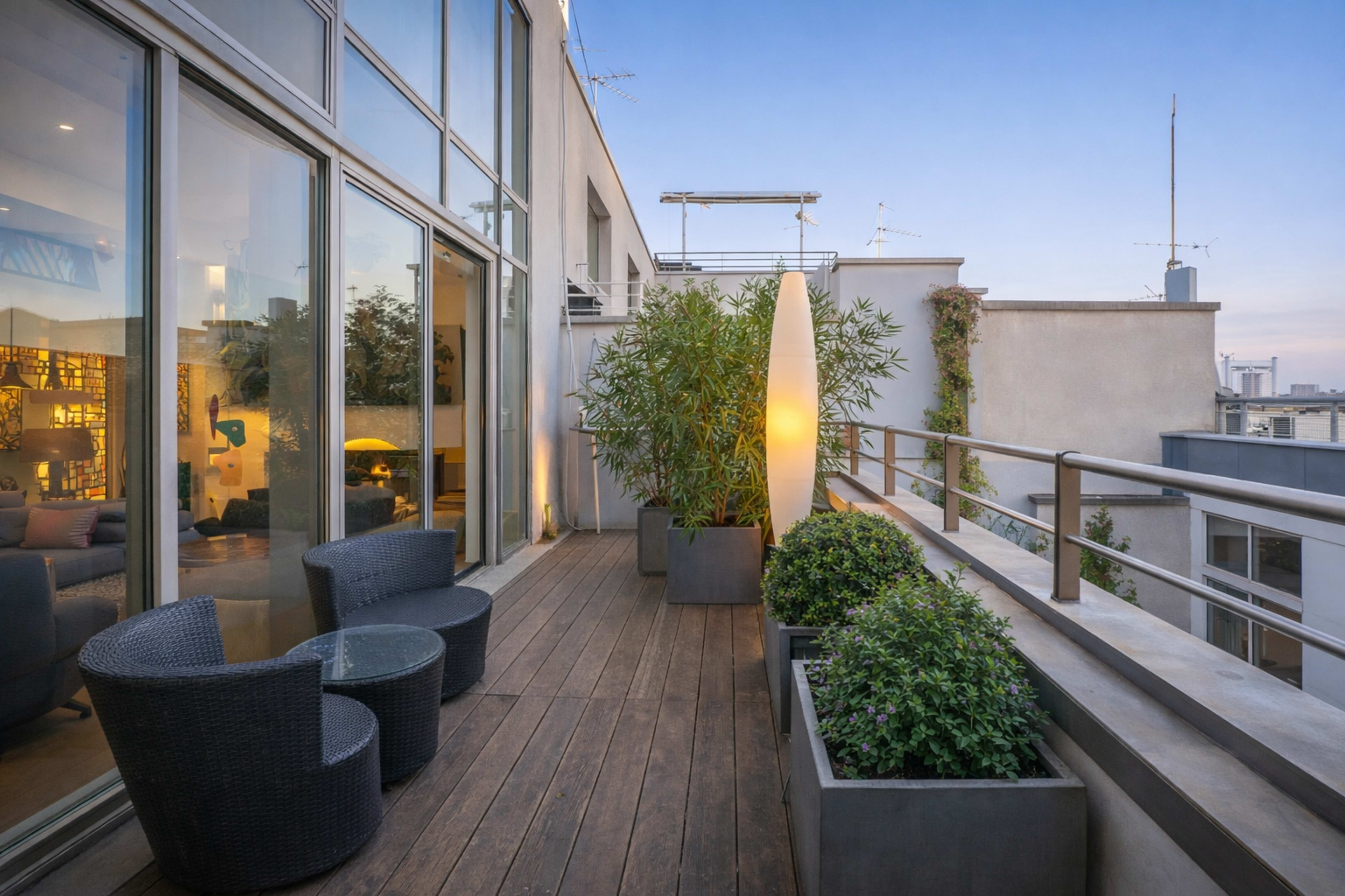 The image shows a rooftop terrace featuring two wicker chairs, a tall decorative light, and potted plants set against a backdrop of large windows and a twilight sky.