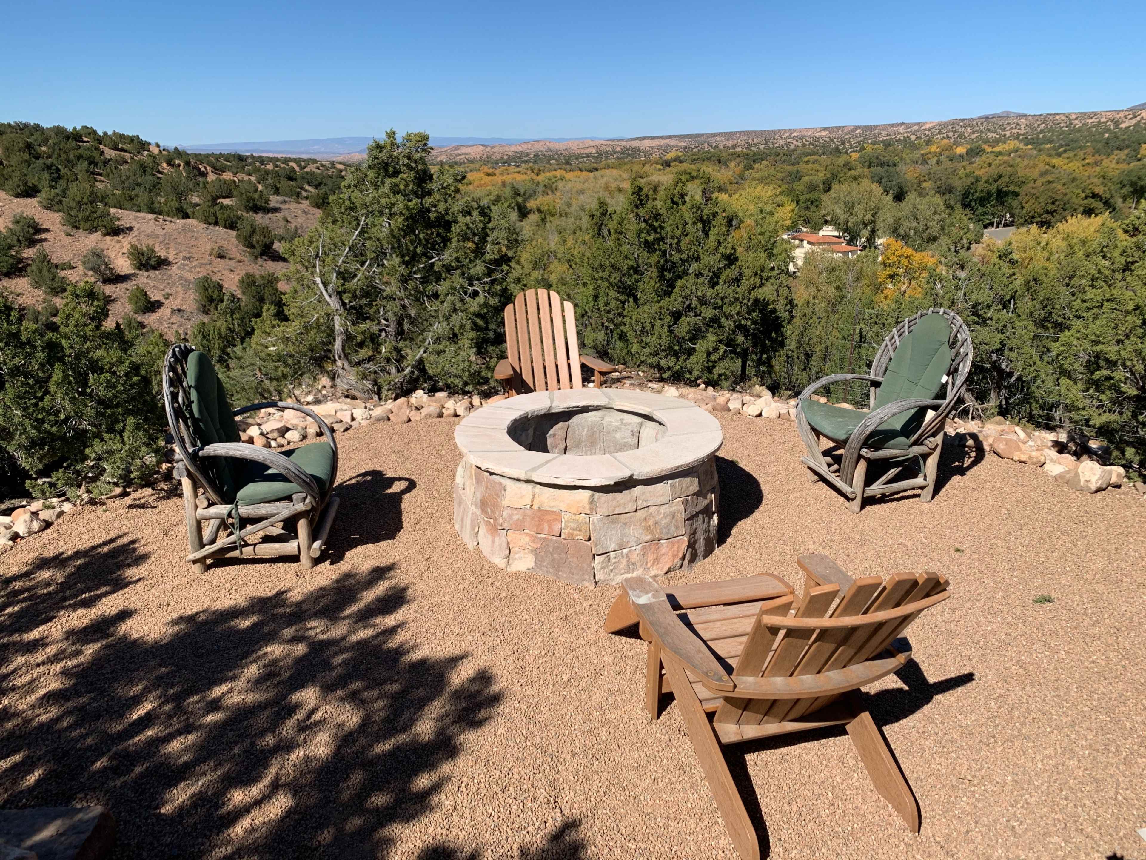A circular stone fire pit surrounded by four wooden chairs on a gravel surface, with a backdrop of rolling hills and trees.