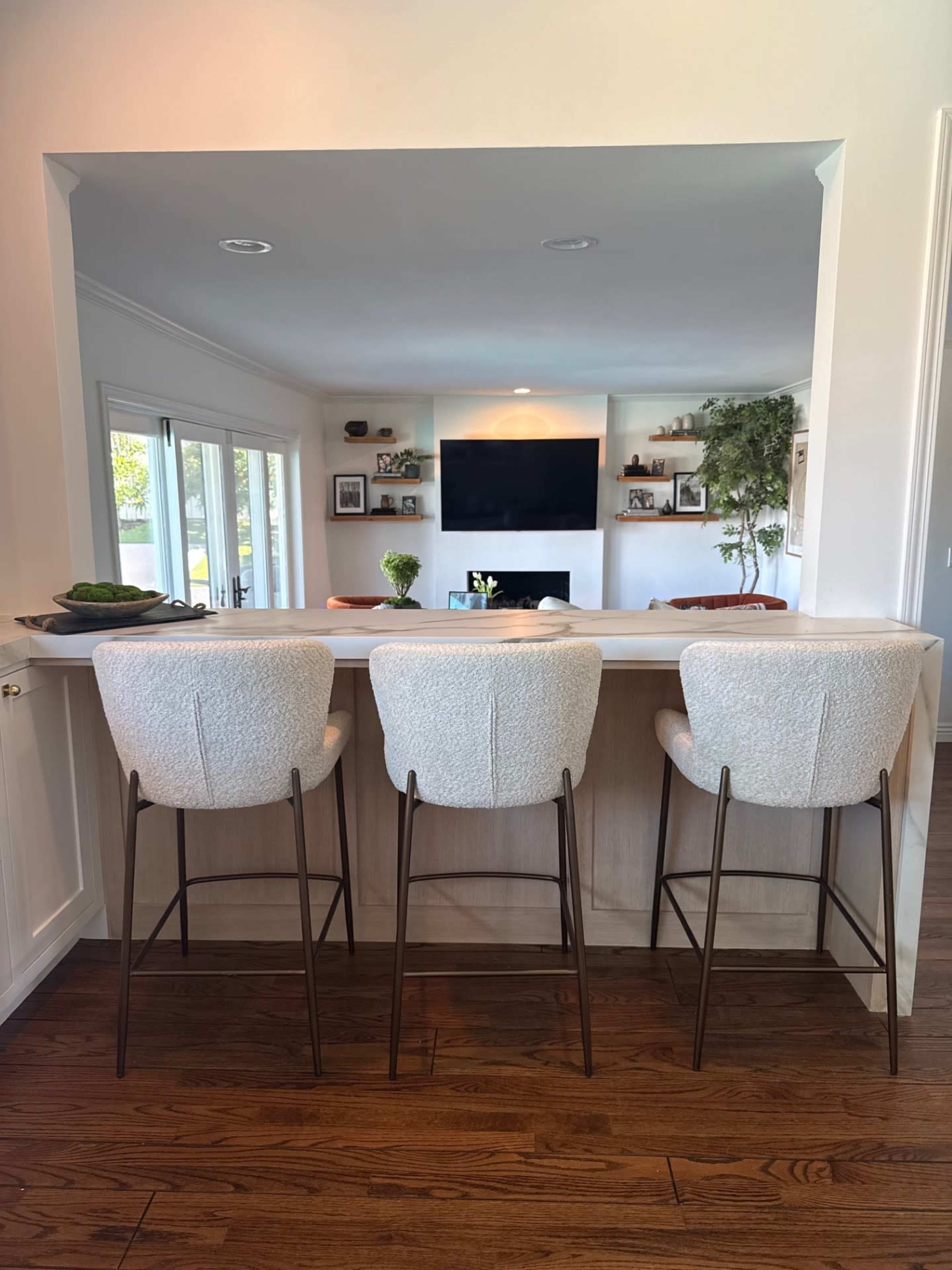 The image shows three modern bar stools with light-colored upholstery situated at a kitchen counter facing a living area with a television and shelves in the background.