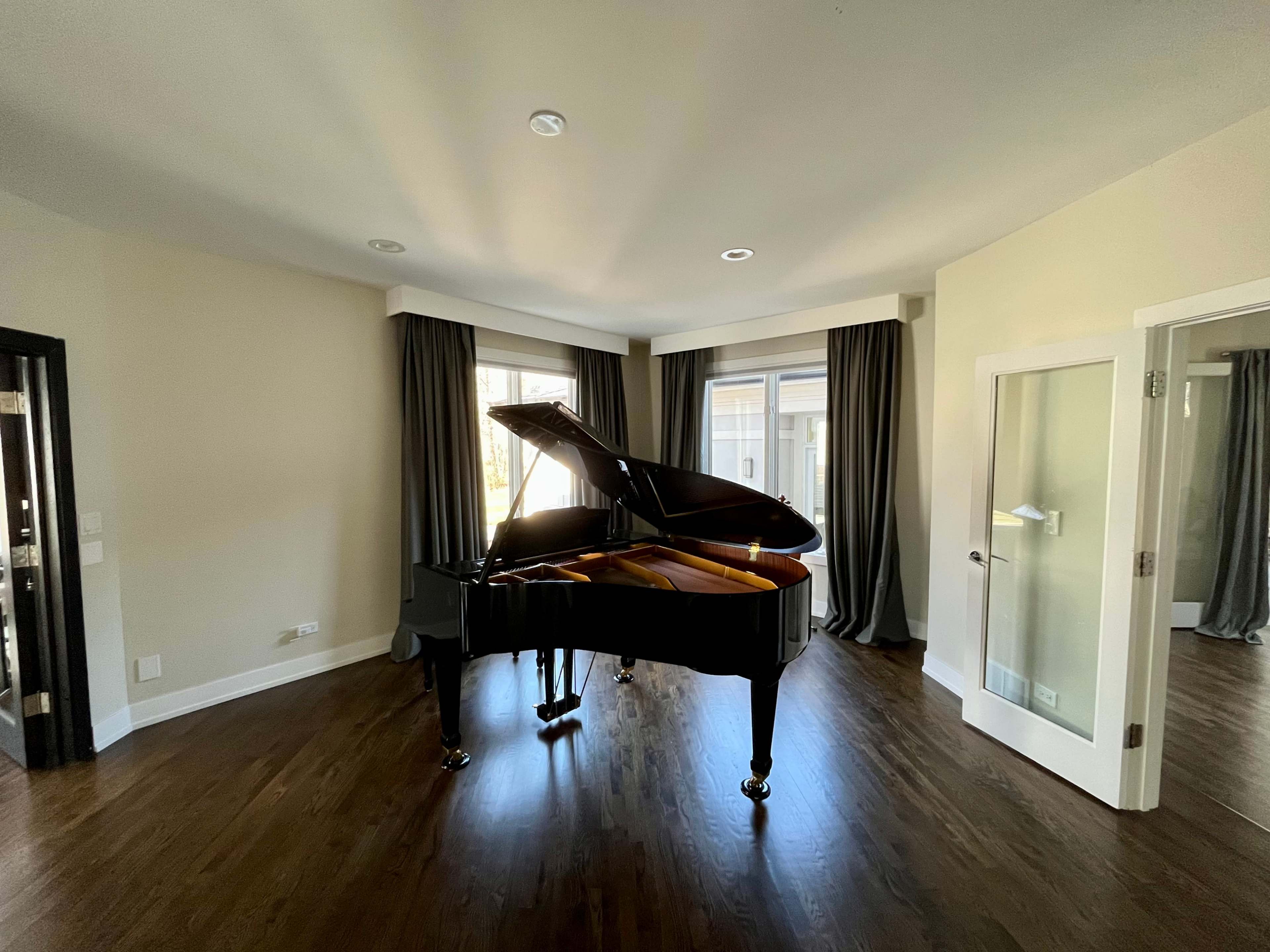 A grand piano is positioned in the center of a well-lit room with large windows and dark curtains.