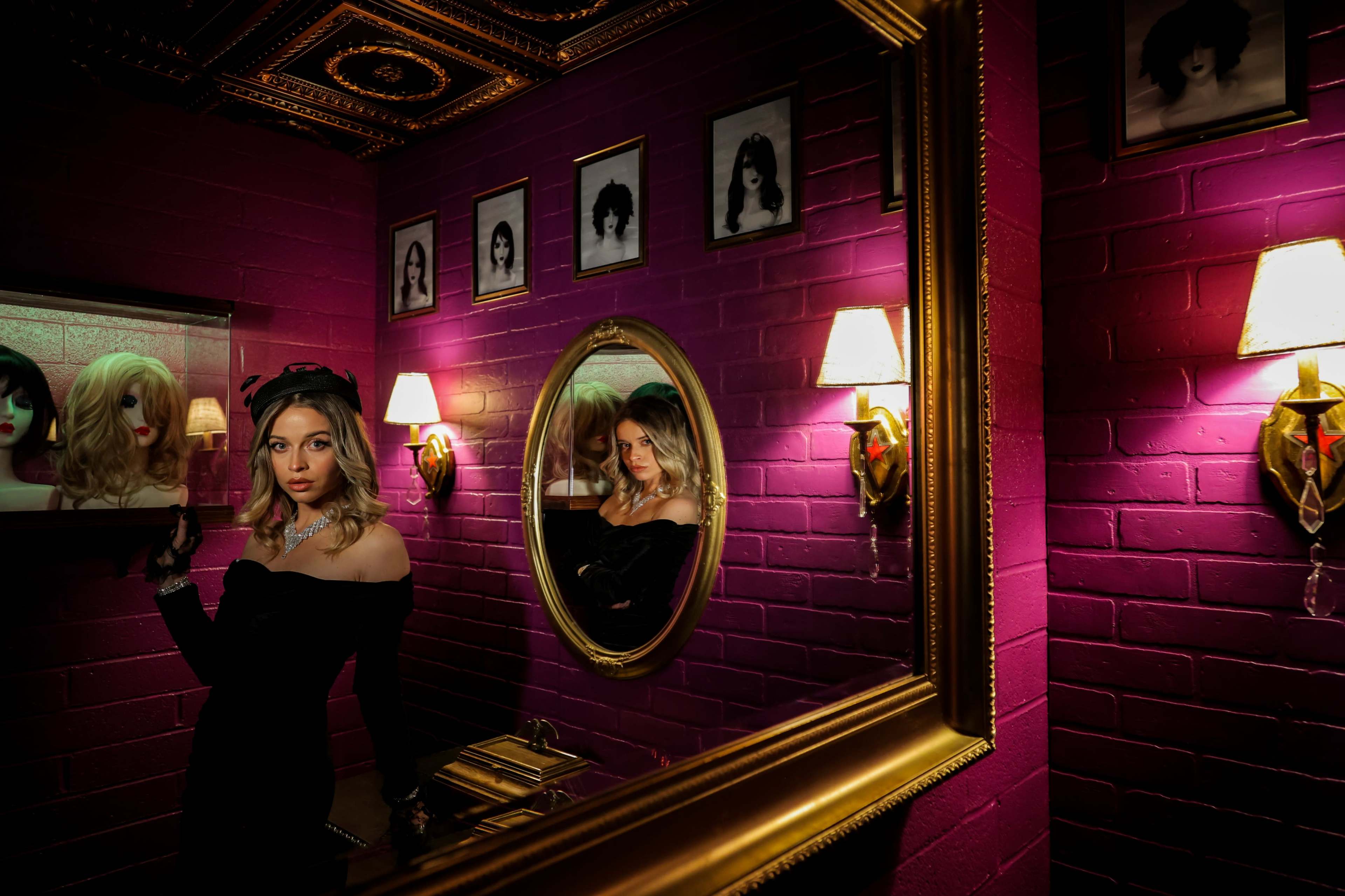 A woman in a black off-shoulder dress stands beside a mirror in a room with purple walls and vintage decor.