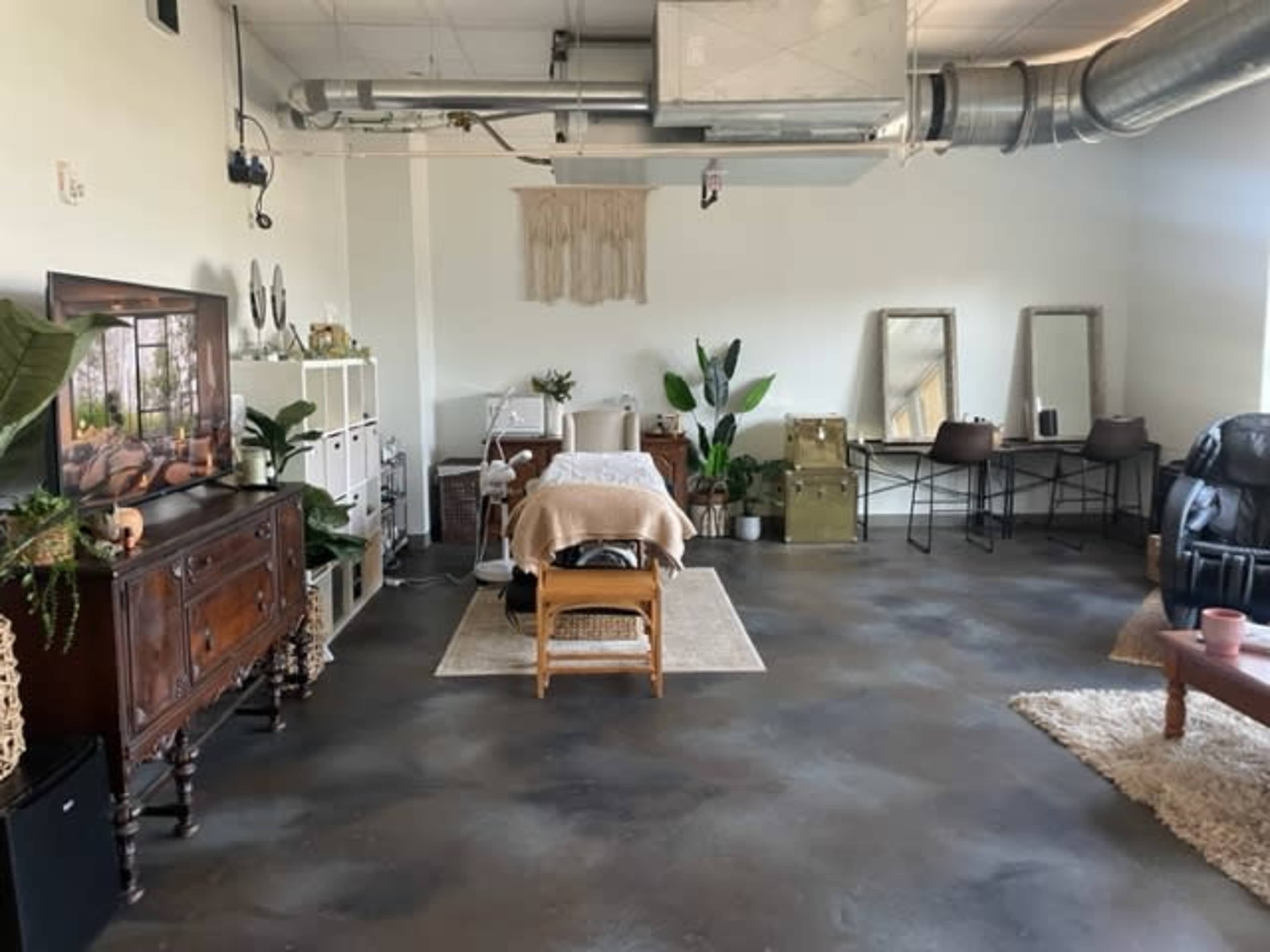A spacious room with a massage table, plants, mirrors, and various furniture including a wooden cabinet and a seating area.