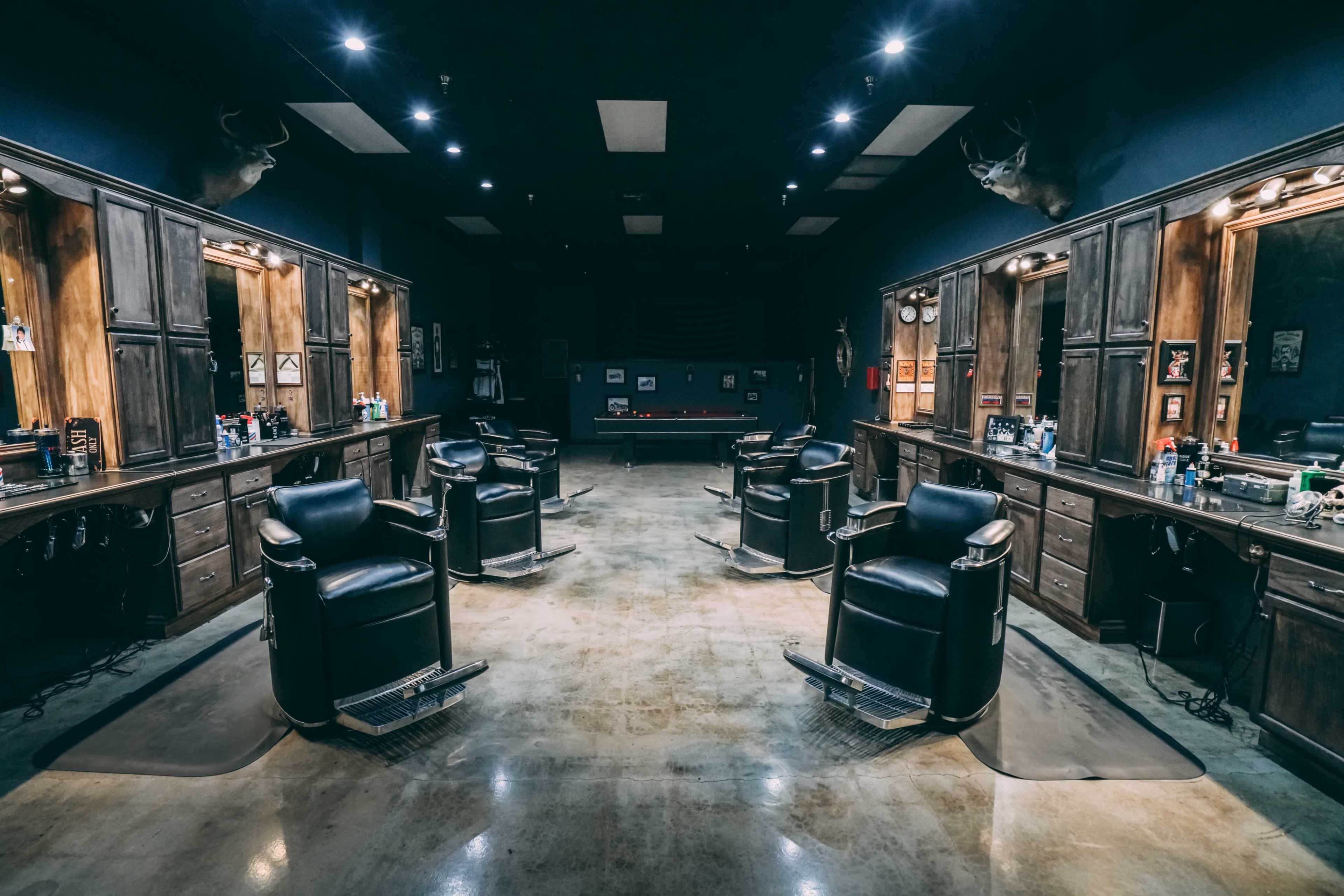A barber shop with four black barber chairs arranged in a spacious, dimly lit room featuring wooden cabinetry and a pool table in the background.