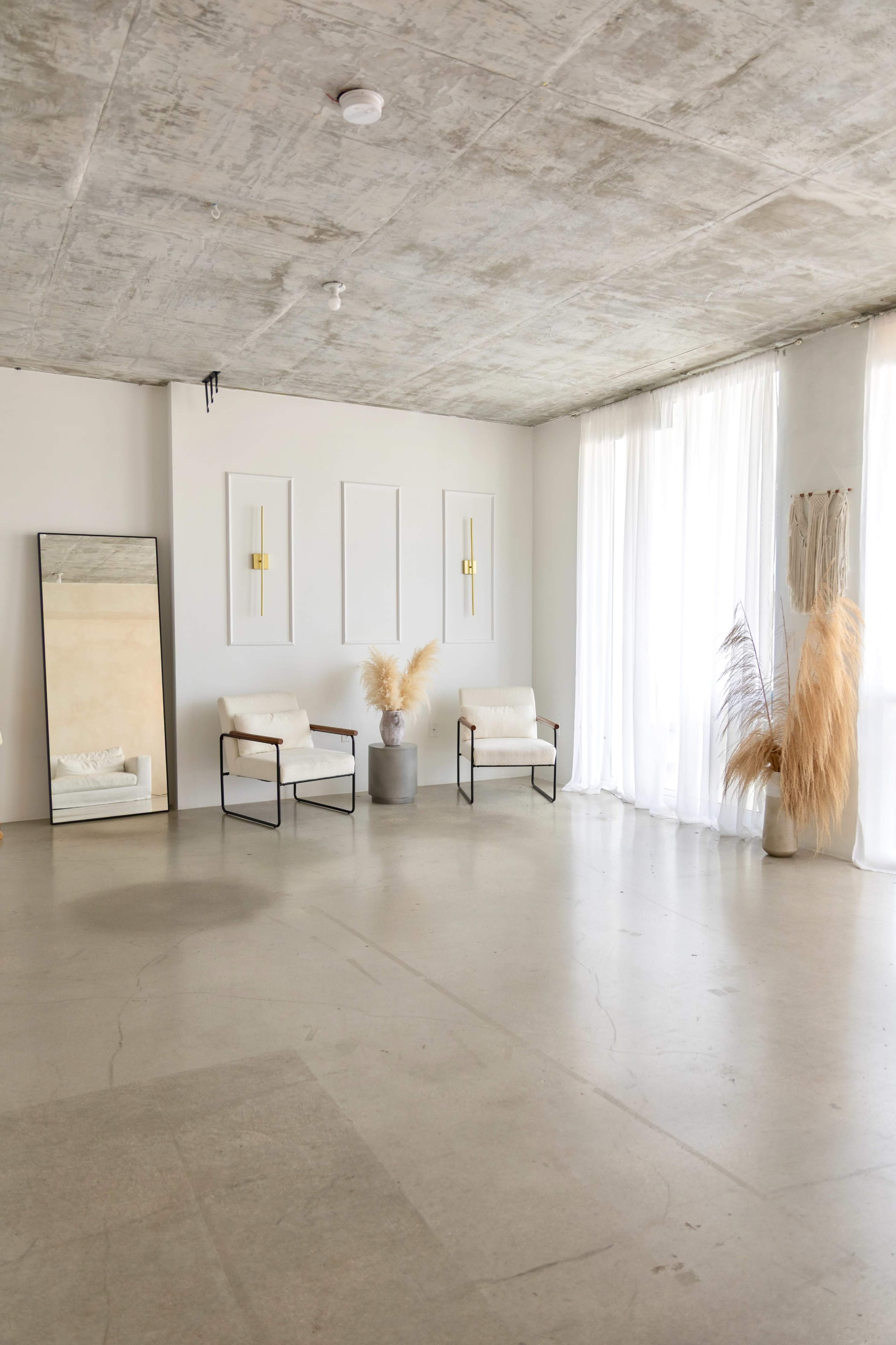 The image features a minimalistic interior space with two armchairs, a large mirror, and decorative plants under natural light from large windows.