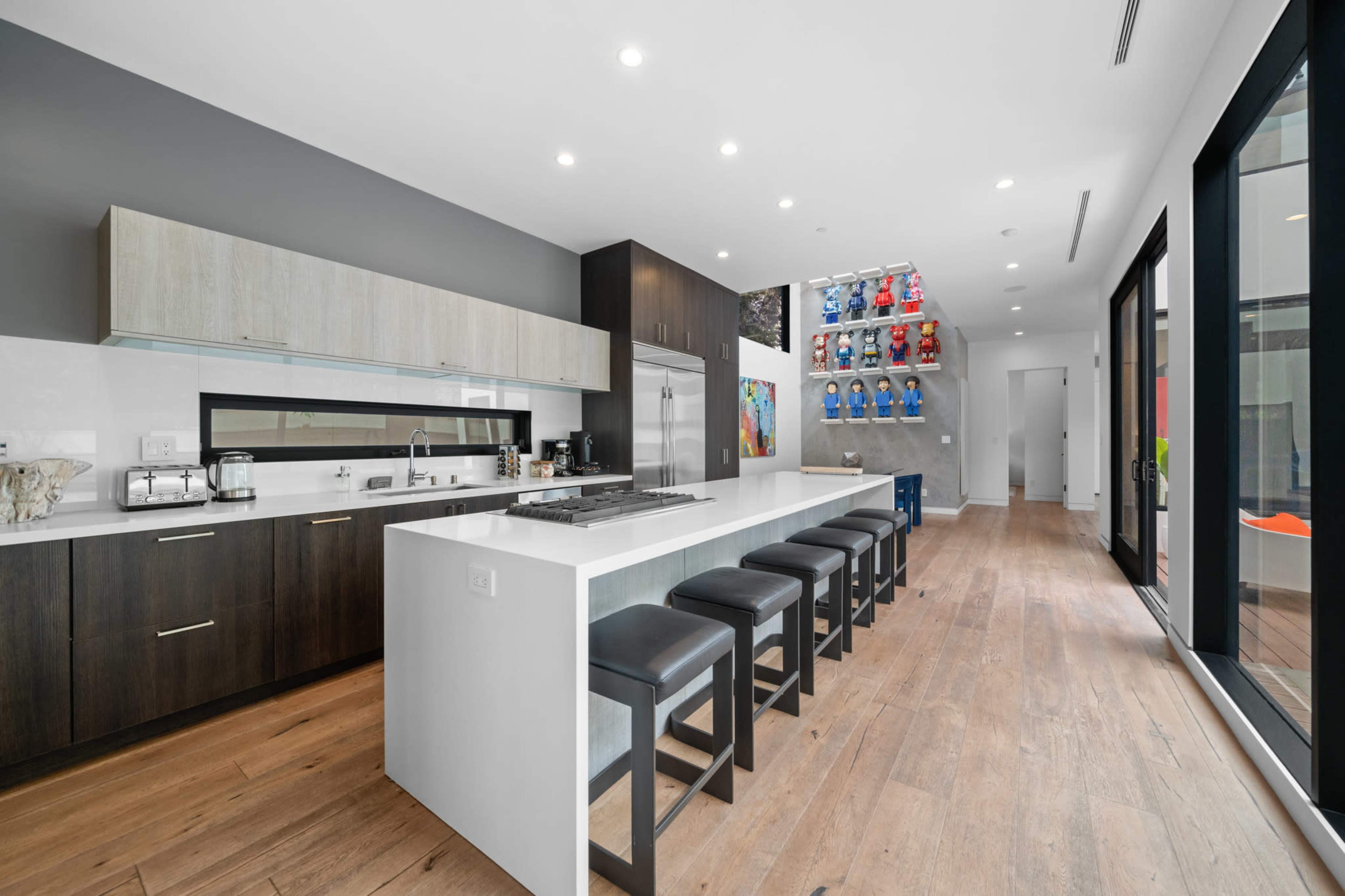 A modern kitchen with sleek cabinetry, stainless steel appliances, and a long island with black stools, illuminated by overhead lights.