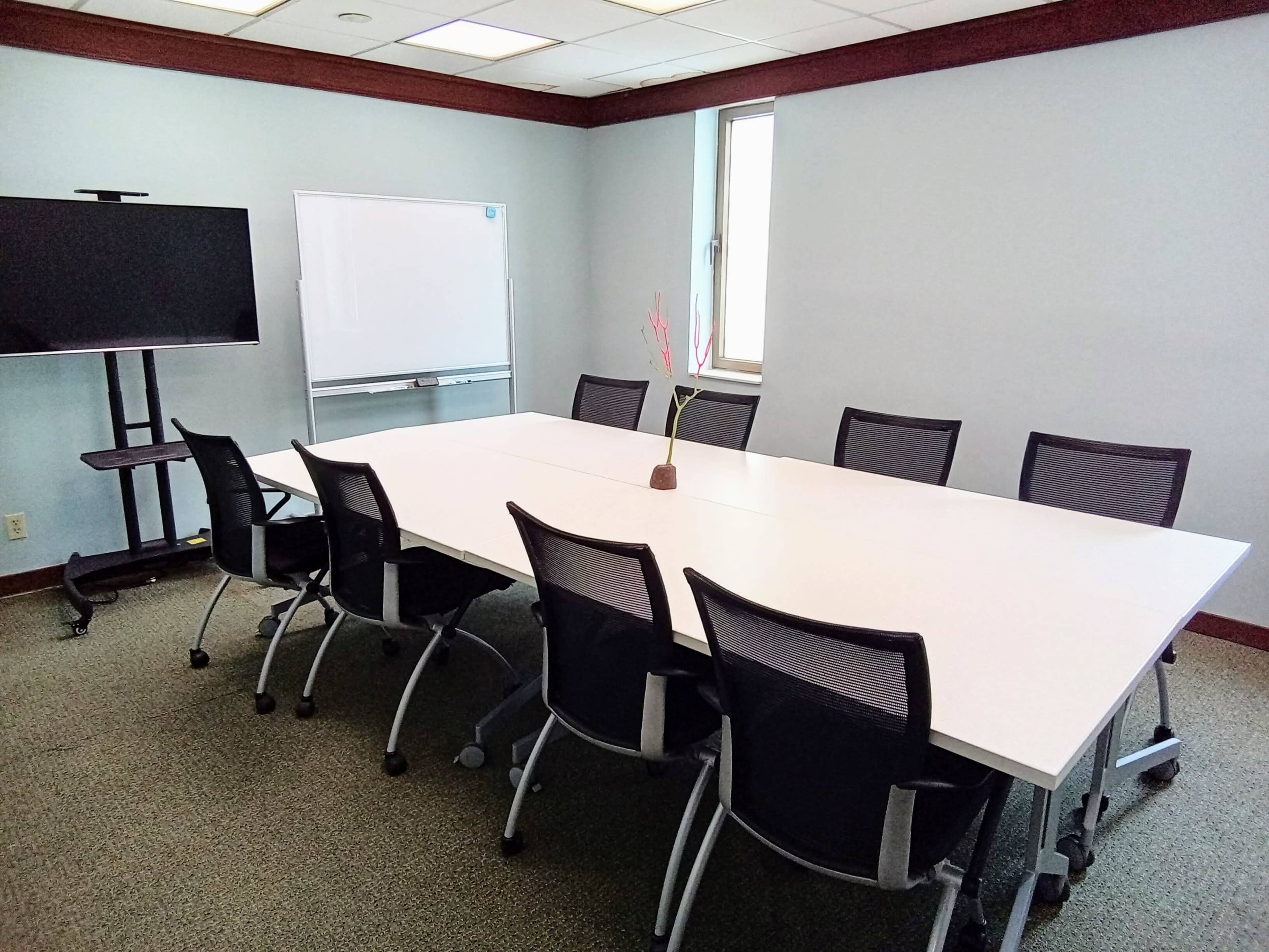 A modern conference room features a large rectangular table surrounded by eight chairs, a television on a stand, and a whiteboard against the wall.