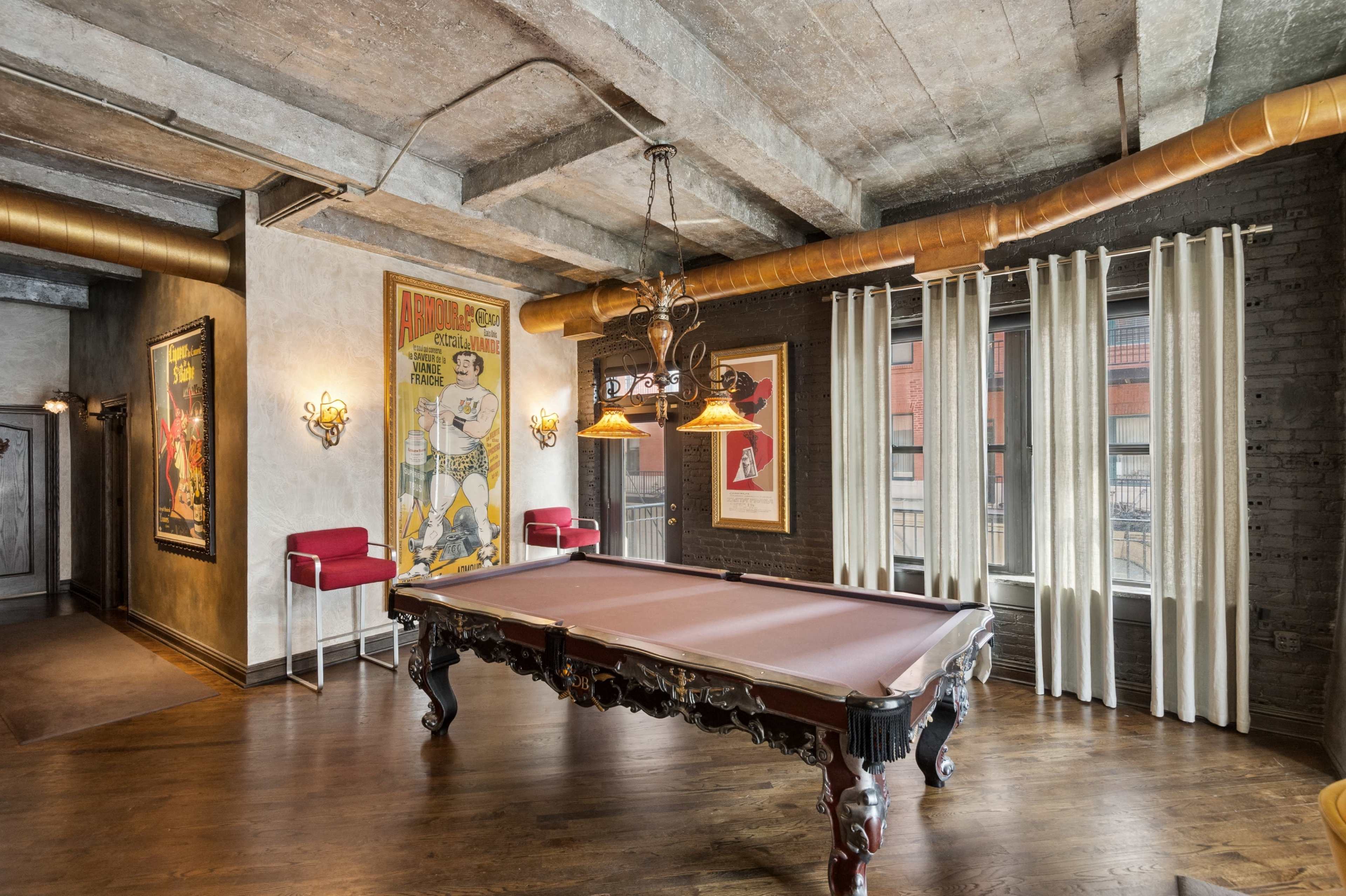 The image shows a game room with a billiard table, exposed concrete ceilings, and large windows, featuring vintage posters and stylish lighting fixtures.