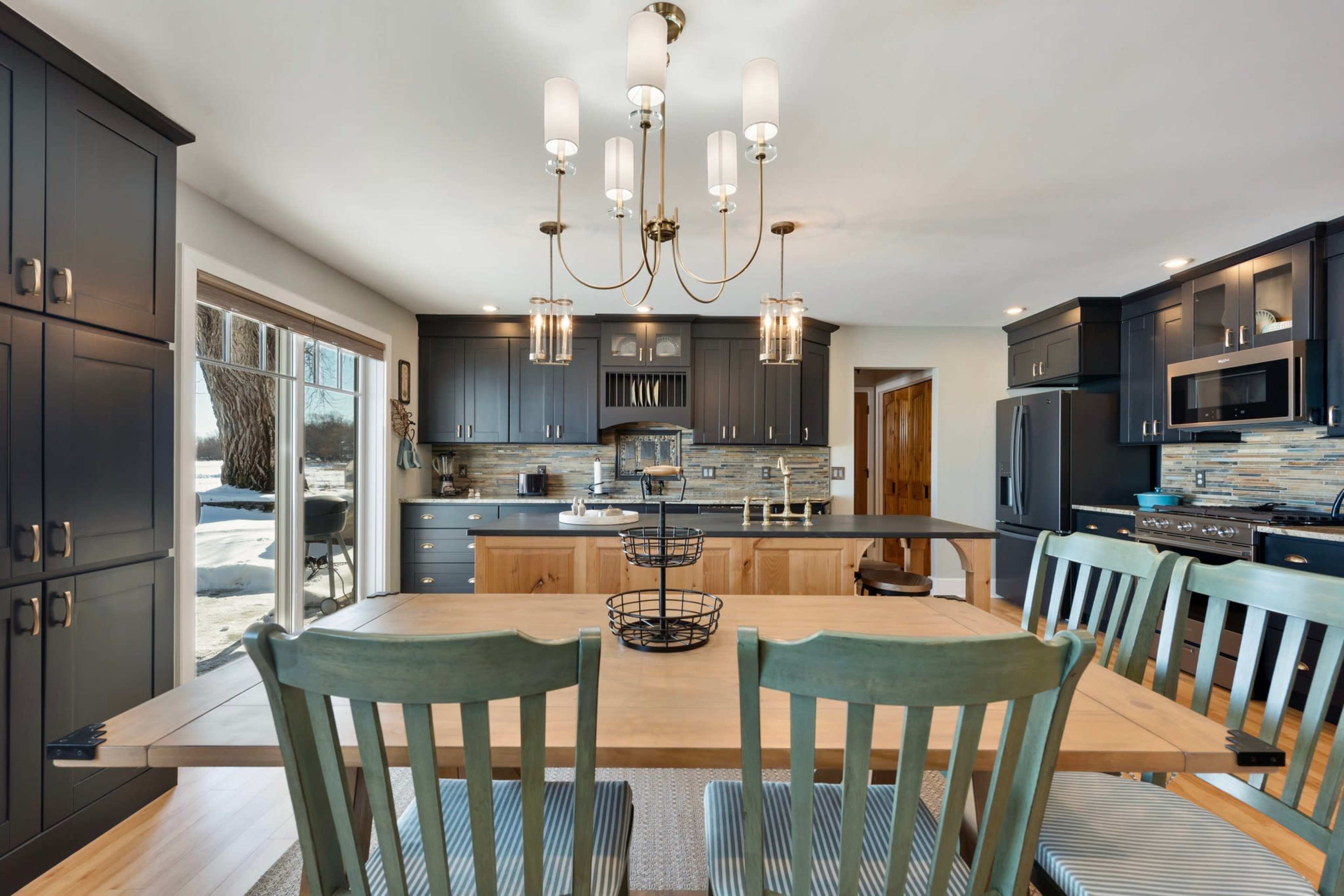 A modern kitchen features dark cabinetry, an island with seating, and a dining area with green chairs under a chandelier.