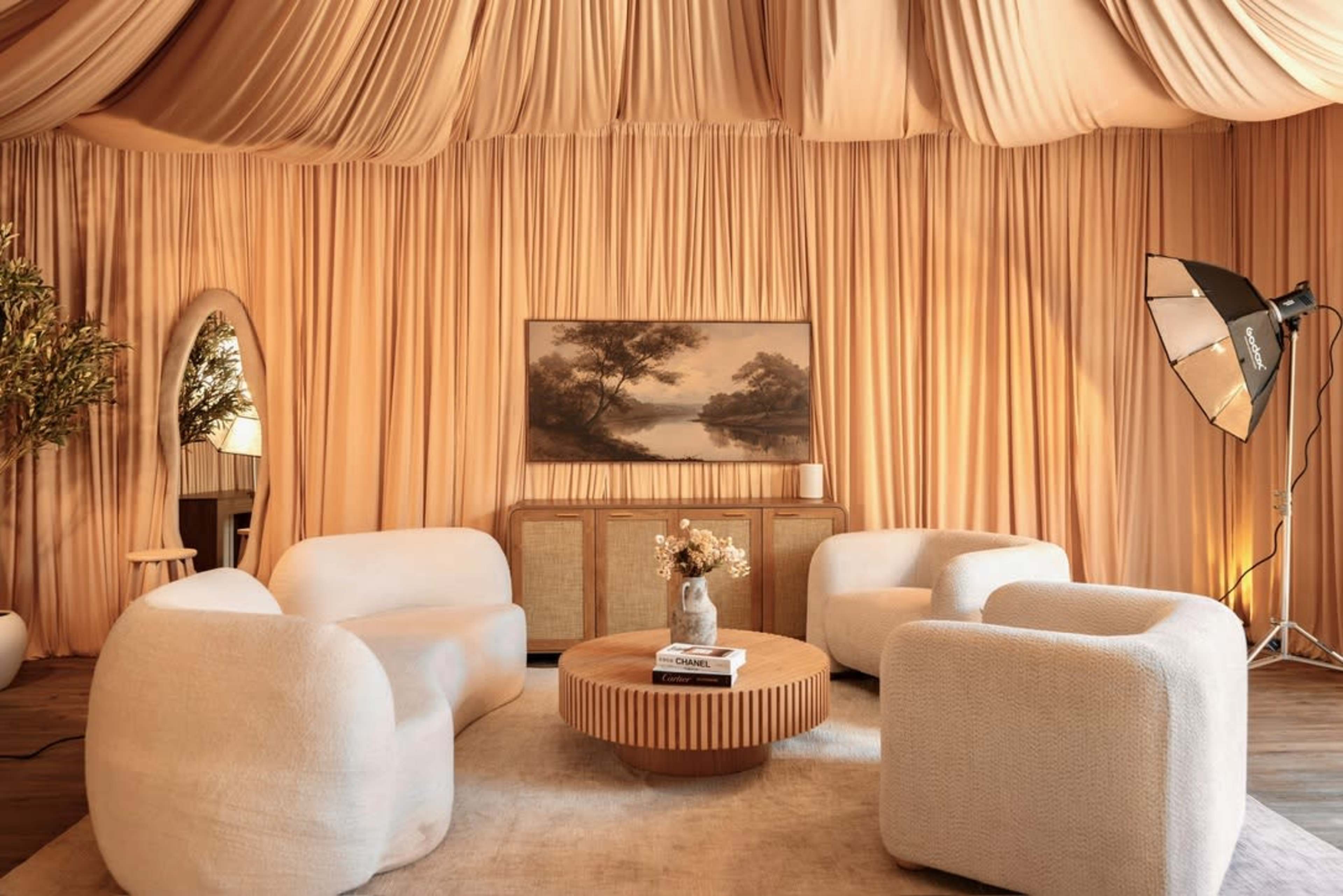 The image shows a cozy living space featuring three rounded chairs around a wooden coffee table, with a large, draped fabric backdrop and a painting on the wall.
