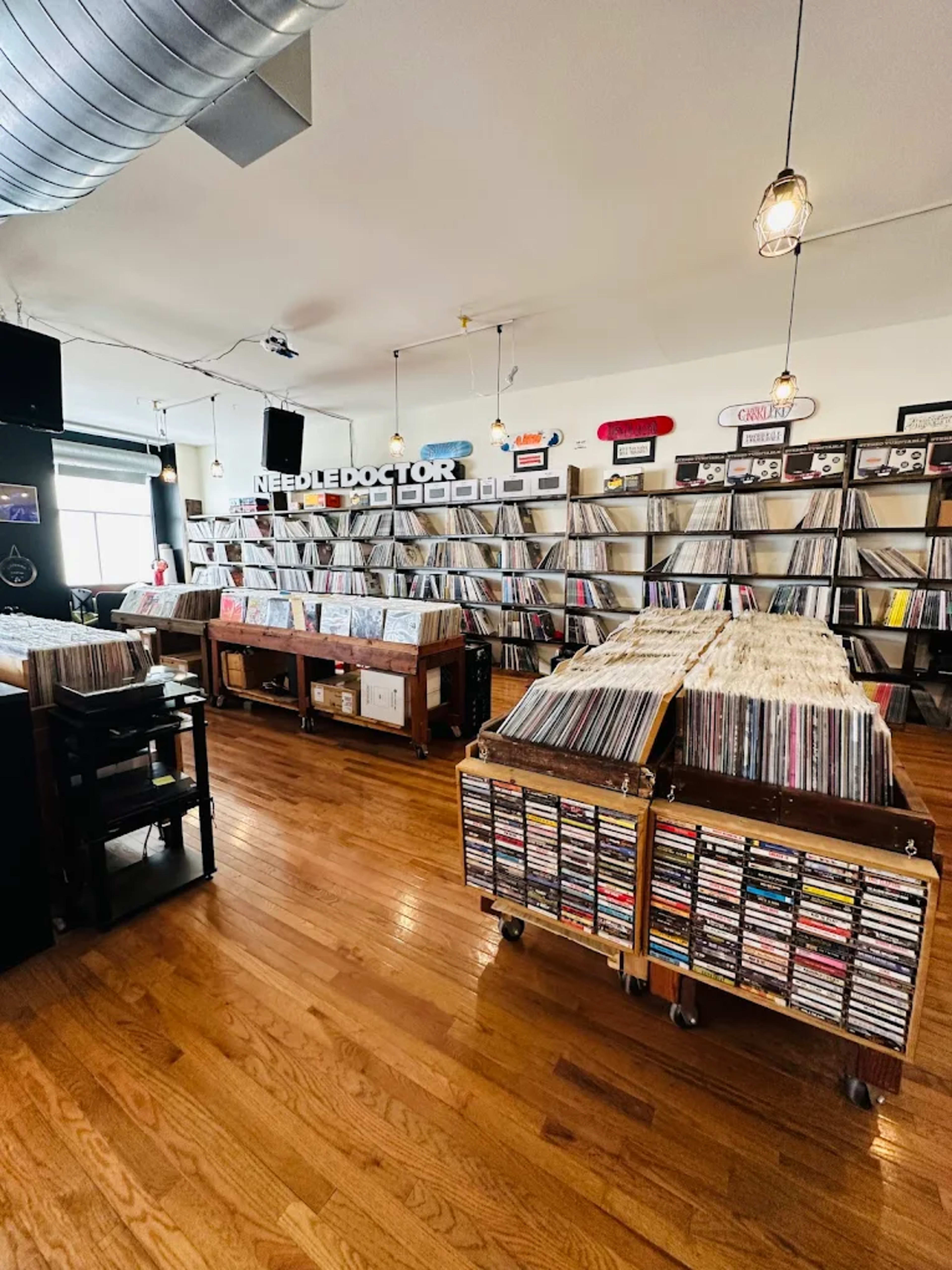 The image shows a vinyl record store with shelves packed with records, wooden carts filled with CDs, and overhead lighting illuminating the space.