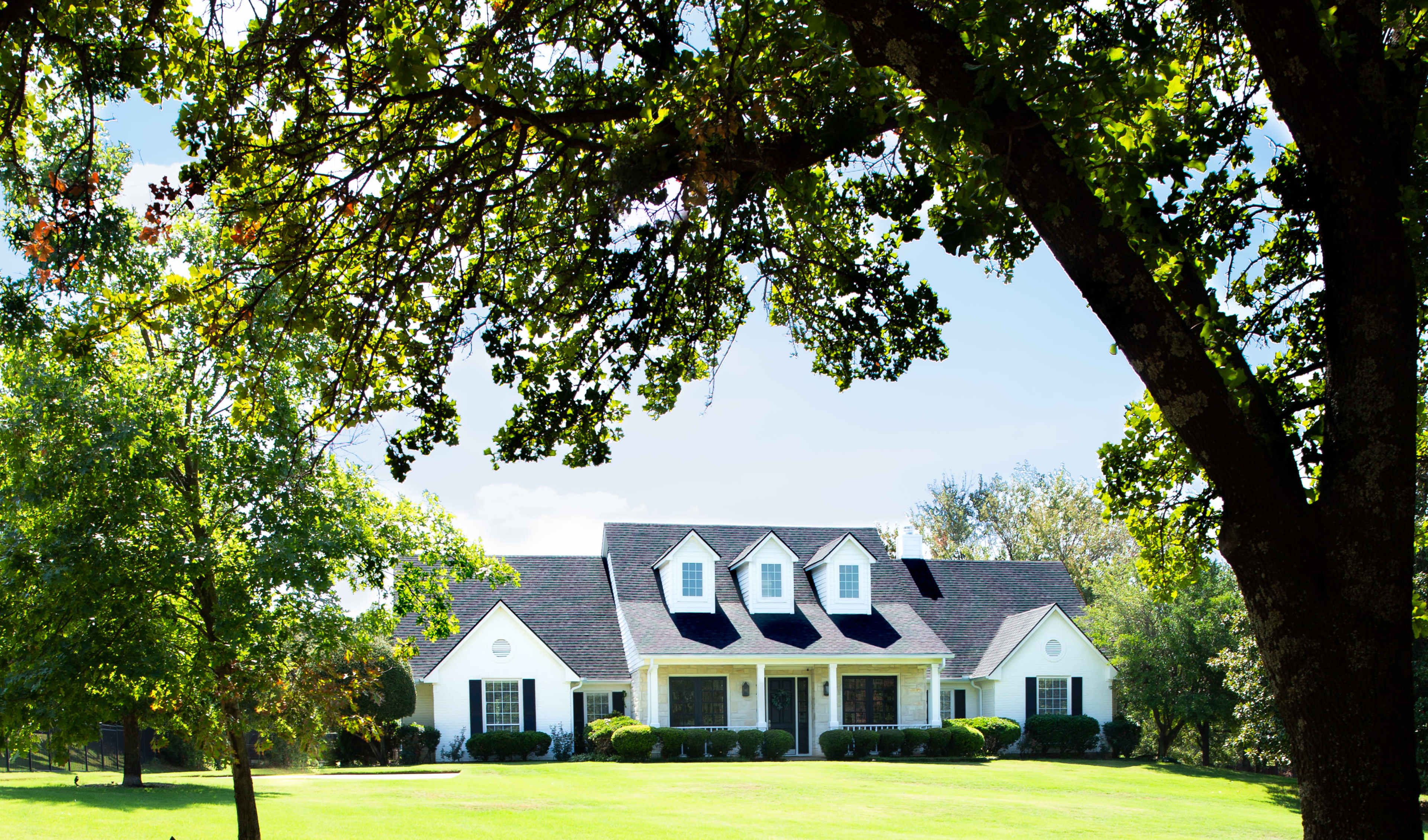 A white house with a sloped roof and multiple gables sits on a green lawn, framed by large trees.